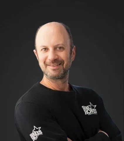 Man with a beard smiling, wearing a black shirt with a logo that says "Star Homes" on the chest and sleeve, against a dark background.