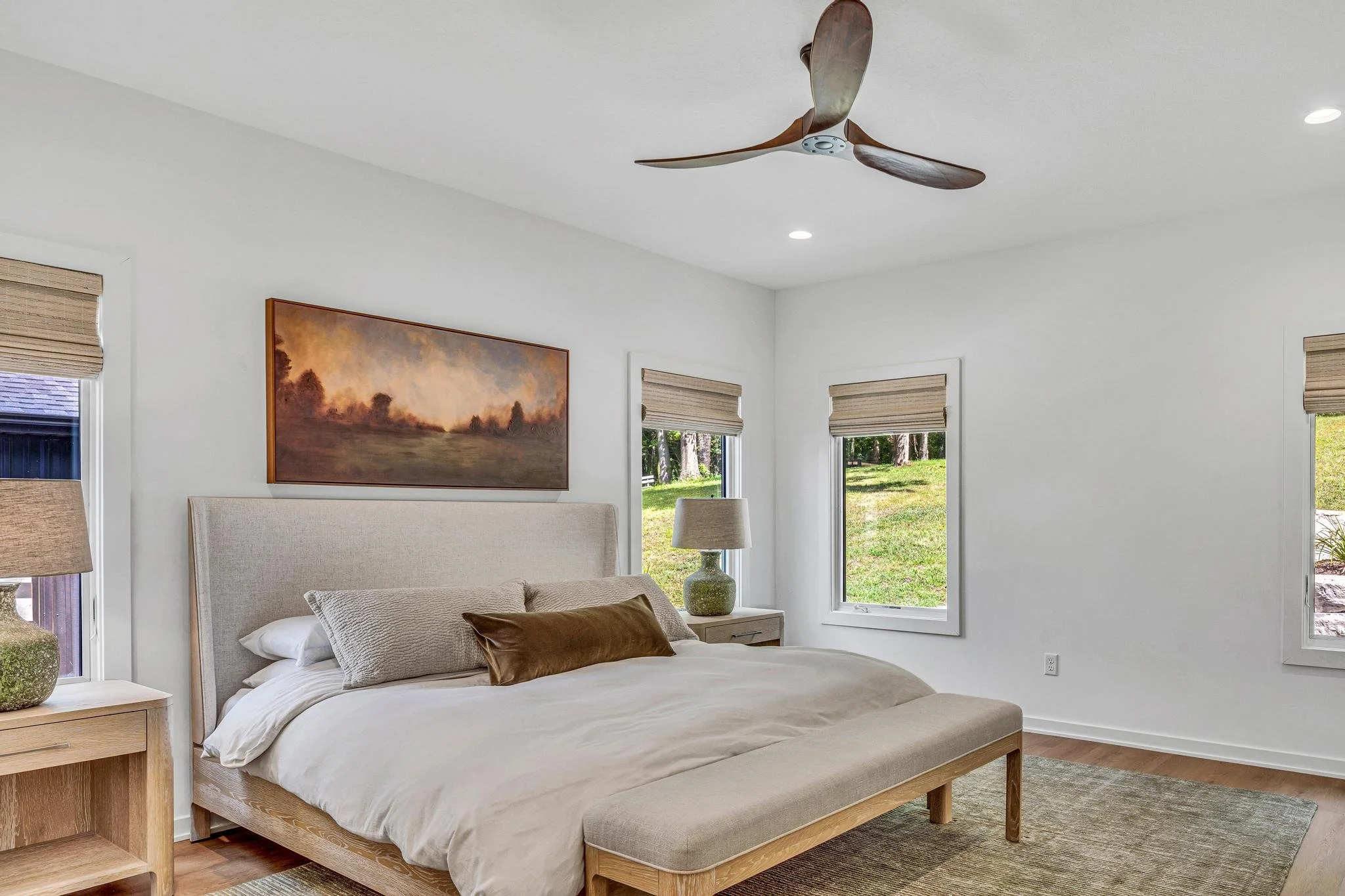 Modern bedroom with white walls, a bed with neutral bedding, wooden nightstands with lamps, a painting above the bed, three windows with beige blinds, a ceiling fan, and a rug on hardwood floor.