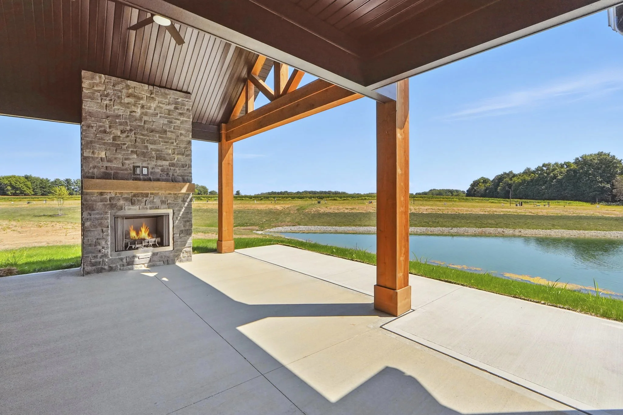 Covered patio with a stone fireplace, wooden beams, and a view of a pond and open field under a clear blue sky.
