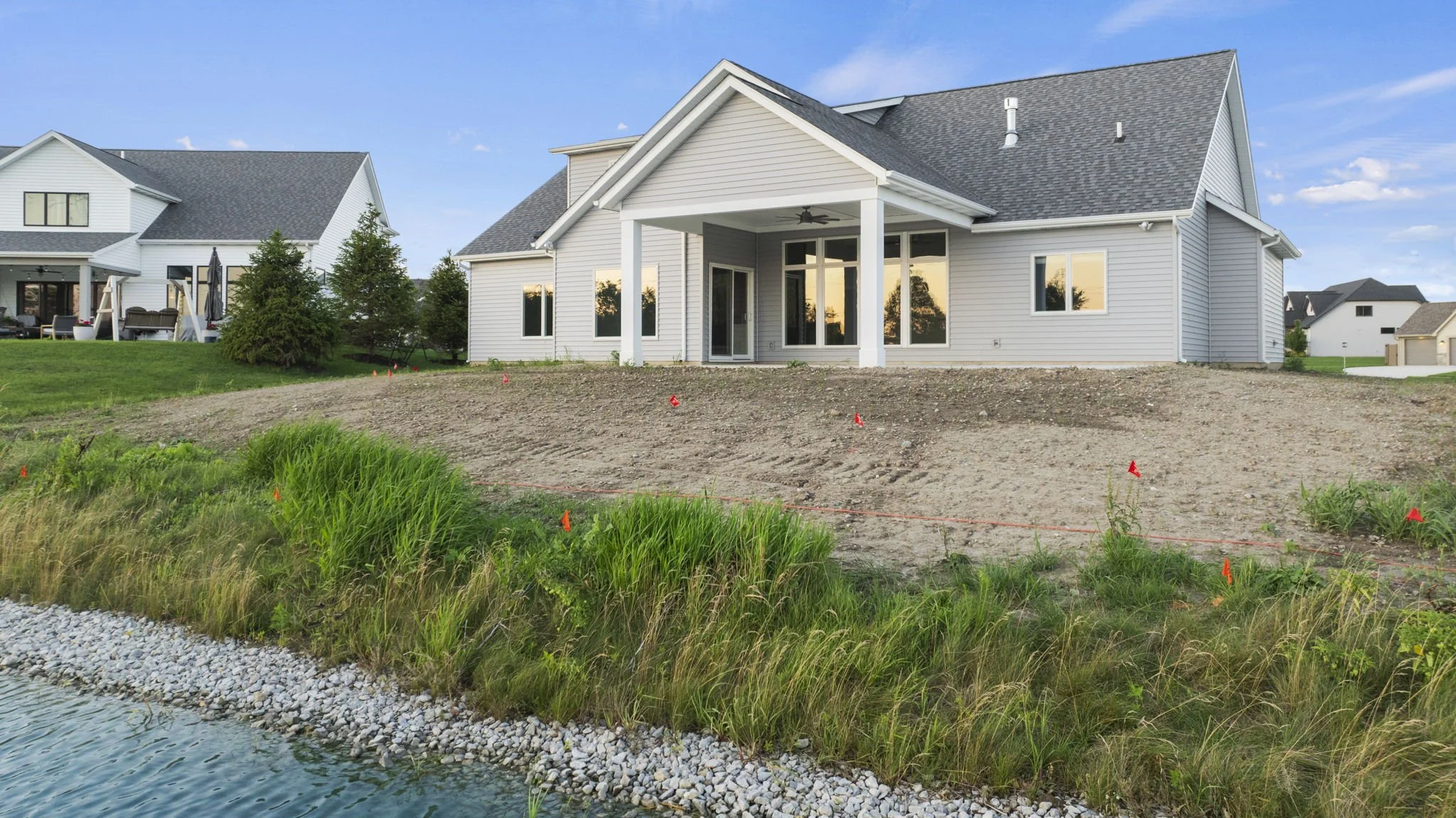 Rear view of a newly constructed house with a covered patio, large windows, and a sloped roof, situated near a body of water with a rocky shoreline and green grass, surrounded by other residential houses.