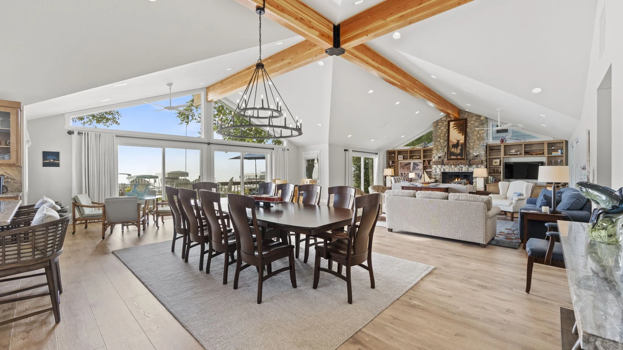 Living room with high vaulted ceiling, large windows, wooden beams, and a mix of modern and rustic furniture, including a fireplace, dining table, and seating area.