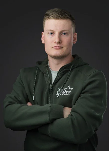 A young man with short, light brown hair and fair complexion posing against a dark background. He is wearing a dark green hoodie with a logo on the chest and has his arms crossed.