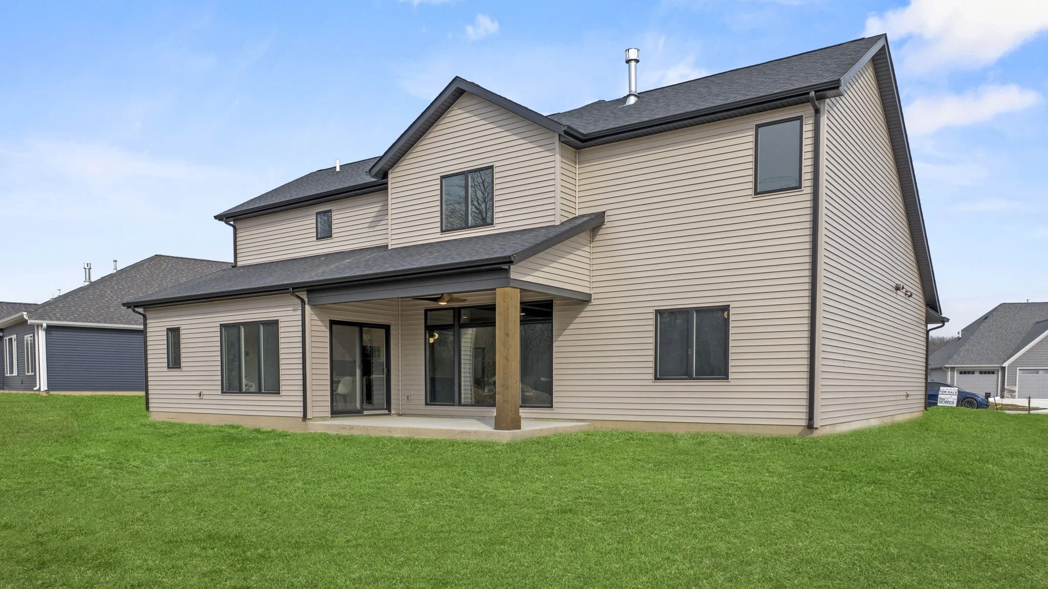 A modern beige two-story house with black window frames, a gray roof, and a covered back patio, surrounded by a green lawn.