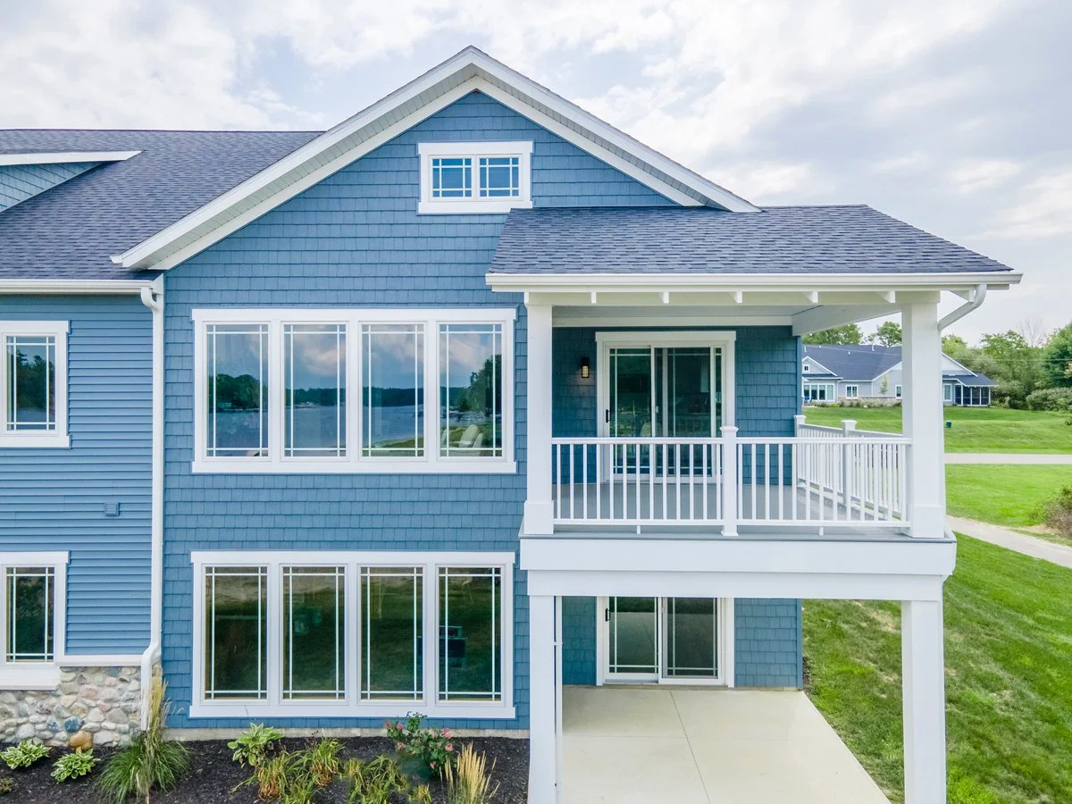 Blue house with white trim, large windows, and a balcony, situated in a green neighborhood with other houses in the background.