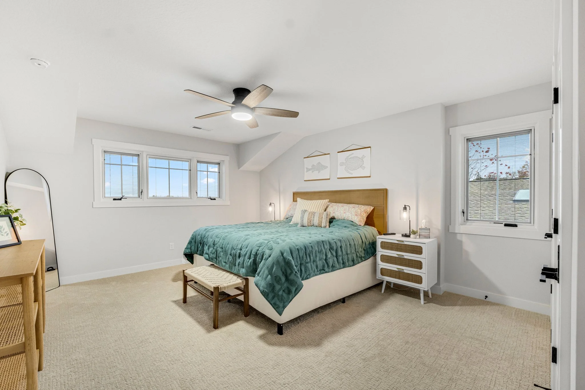 A bedroom with a white bed, teal comforter, and throw pillows, wooden headboard, two nightstands with lamps, a small bench at the foot of the bed, a window with blinds, and a ceiling fan with light.