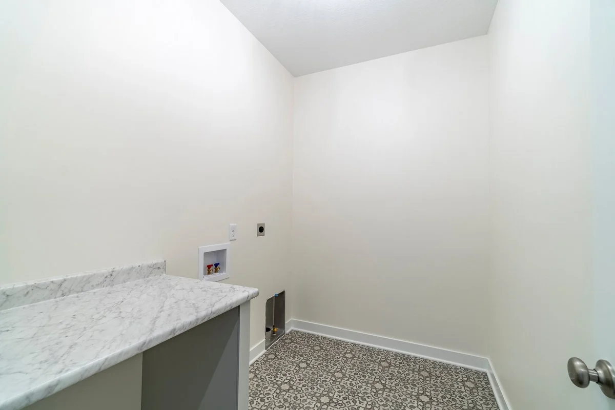 Empty laundry room with white walls, a drain, and water hookup on the wall, a granite countertop on a cabinet, and patterned tile flooring.