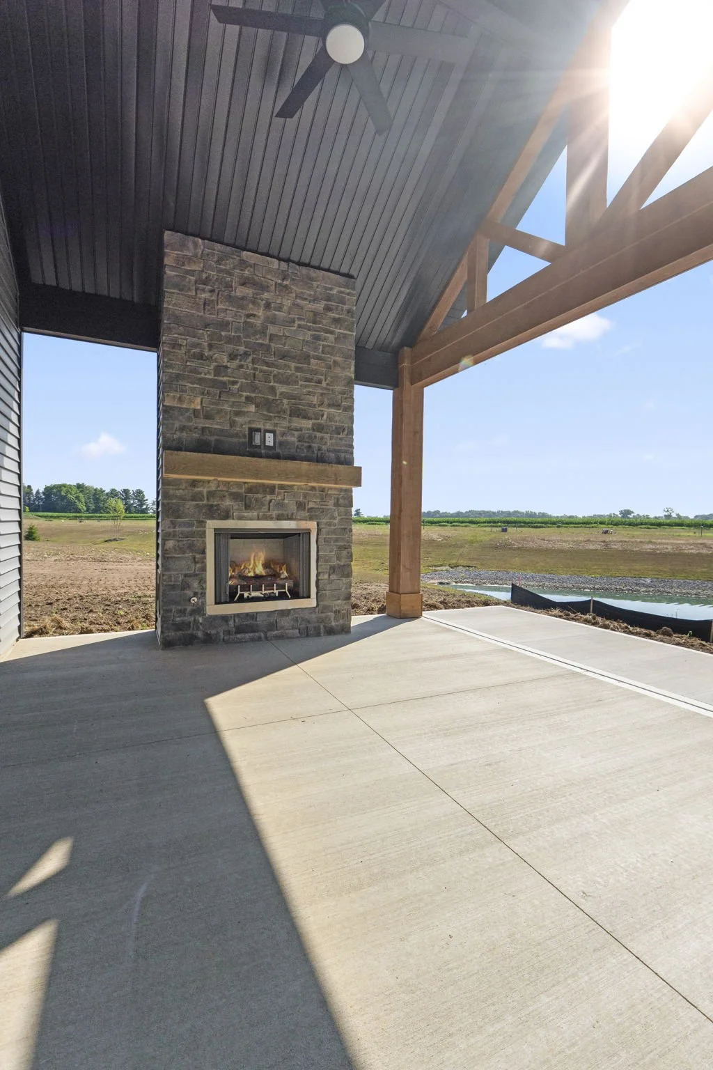 Exterior patio with concrete floor, stone fireplace, wooden beams, ceiling fan, open sky, and a vast open field in the background.
