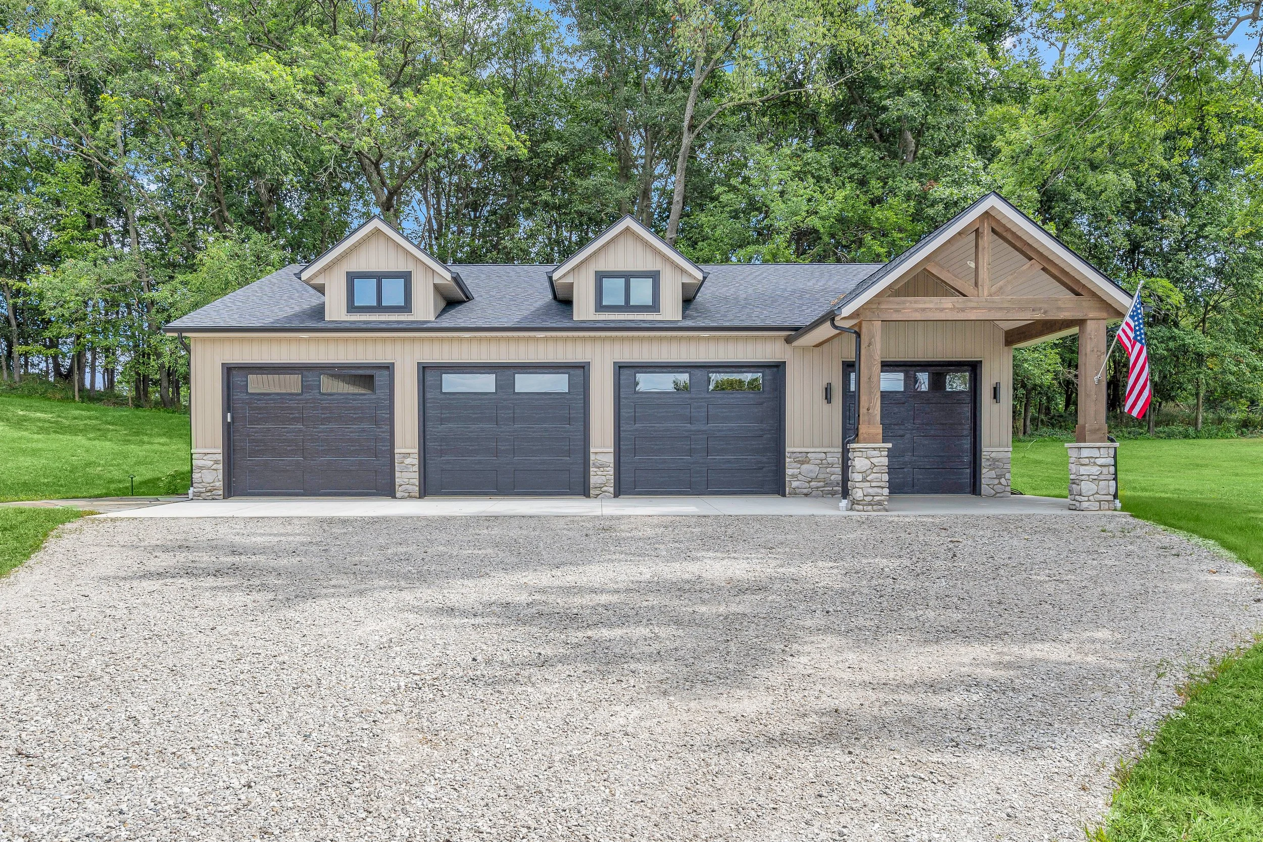 A modern garage with four black doors, stone accents at the bottom, beige siding, a covered porch area with wooden beams, an American flag hanging on the porch, and surrounded by green trees and grass.