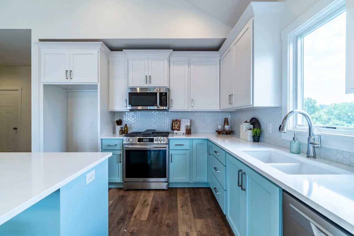 Modern kitchen with white upper cabinets and light blue lower cabinets, stainless steel appliances including a microwave and oven, a large window above the sink, and hardwood flooring.