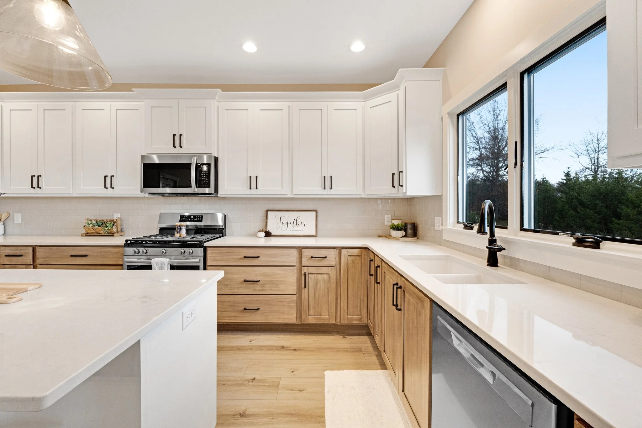 Modern kitchen with white upper cabinets, natural wood lower cabinets, a large window over the sink, stainless steel appliances, and light-colored countertops.
