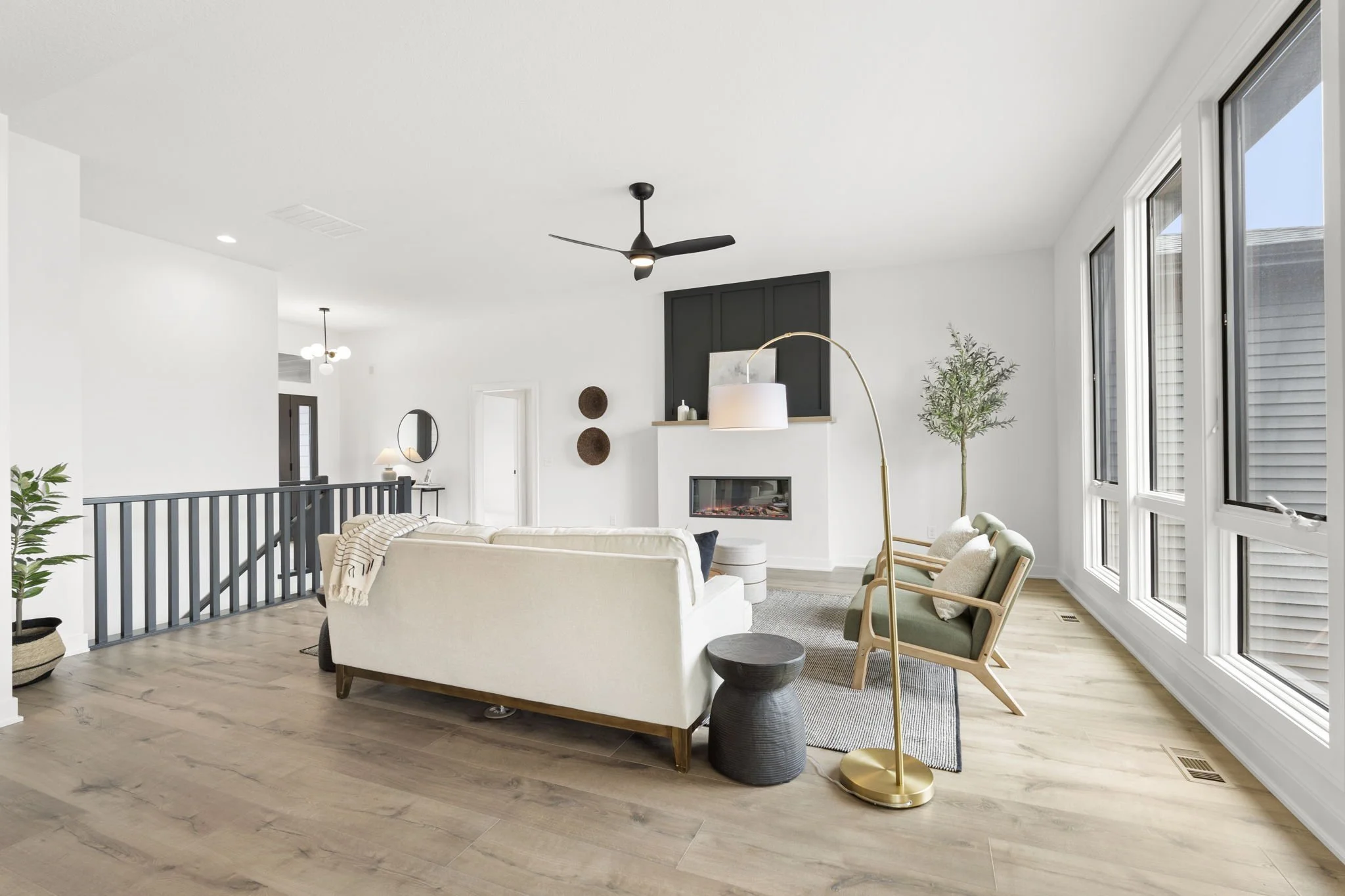 Living room with white walls, wooden floor, large windows, cream sofa, two green armchairs, black ceiling fan, black fireplace, a white standing lamp, and plants.