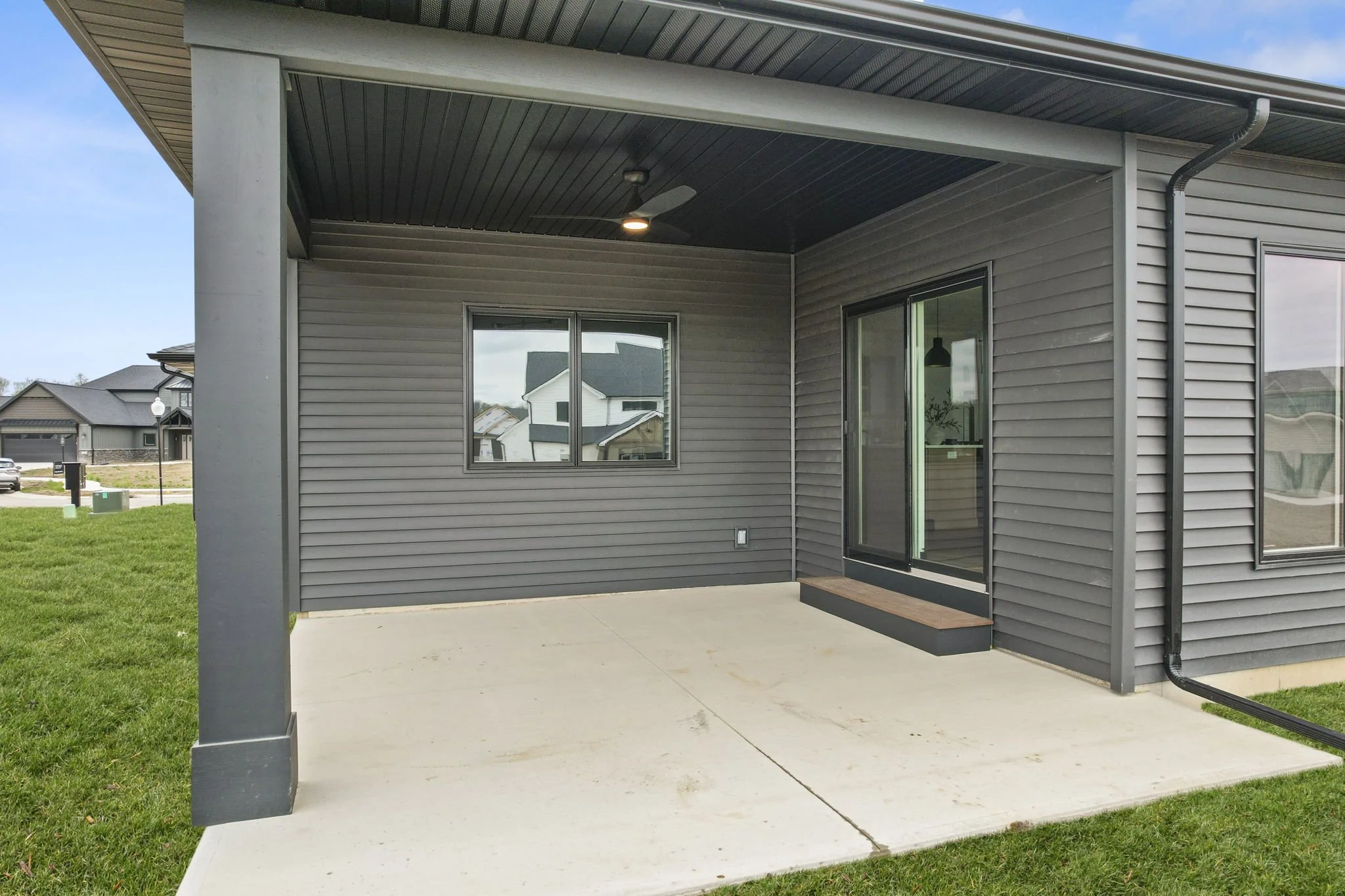 Back patio of a modern house with gray siding, a concrete slab floor, sliding glass door, and a window, under a covered roof with a ceiling fan and lights.