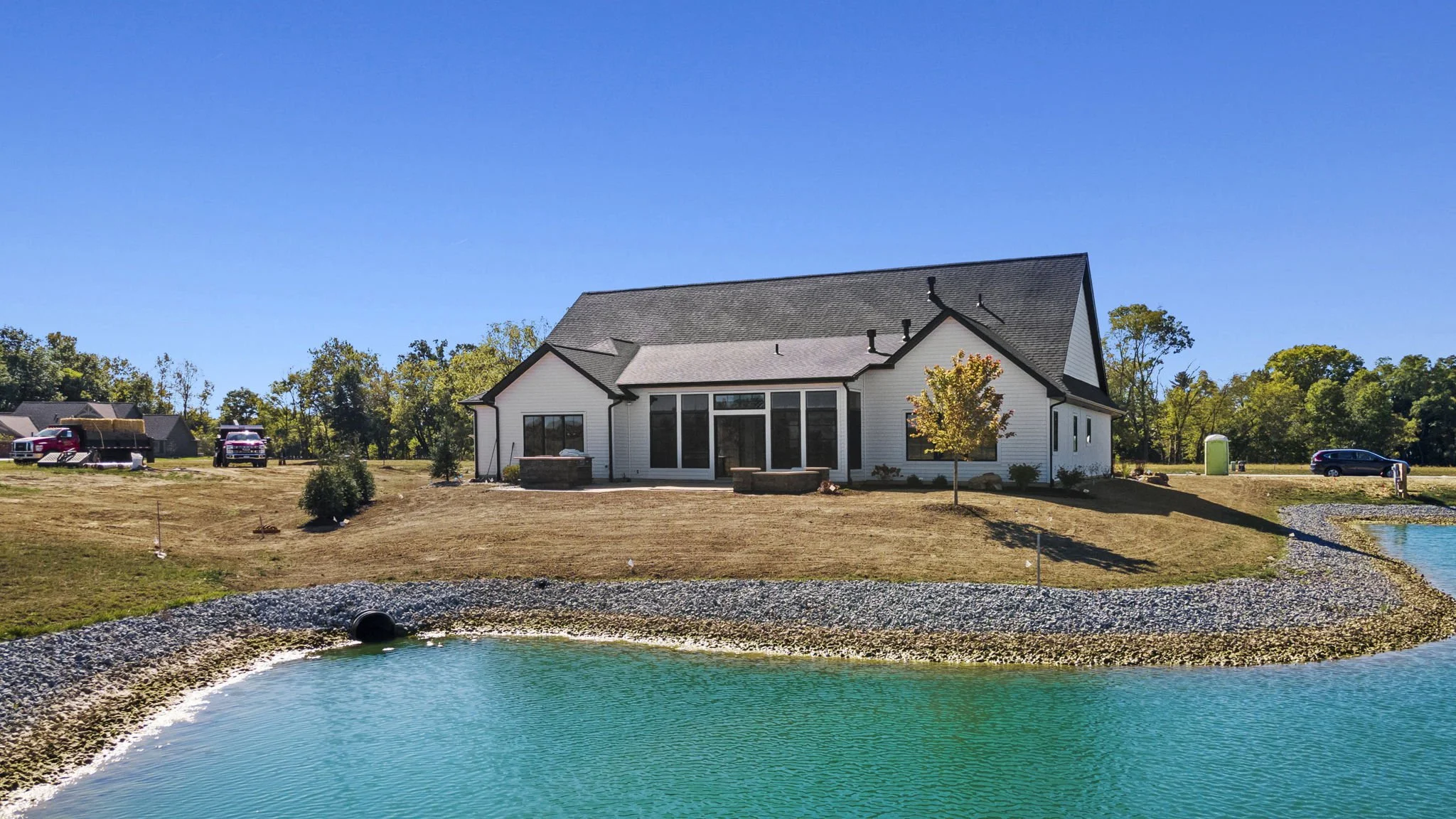 A modern house with a dark gray roof, white exterior walls, and large windows, situated on a sloped grassy lawn next to a pond with a curved shoreline of rocks, under a clear blue sky. There are trees and some vehicles in the background.