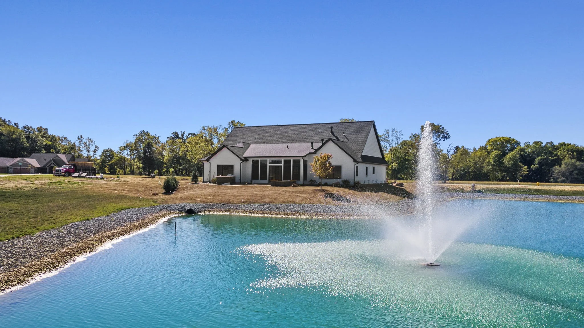 View of a white house with a dark roof across a pond with a tall fountain, outdoors on a sunny day with a clear blue sky.