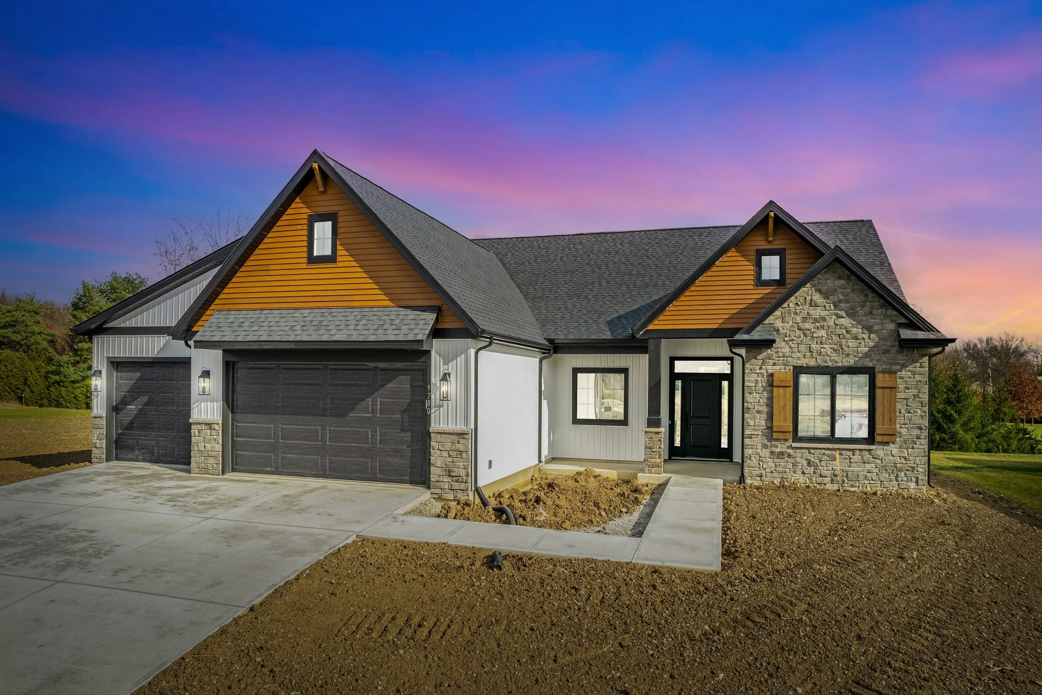 Newly constructed modern house with dual garage doors, stone and siding exterior, and a colorful sunset sky