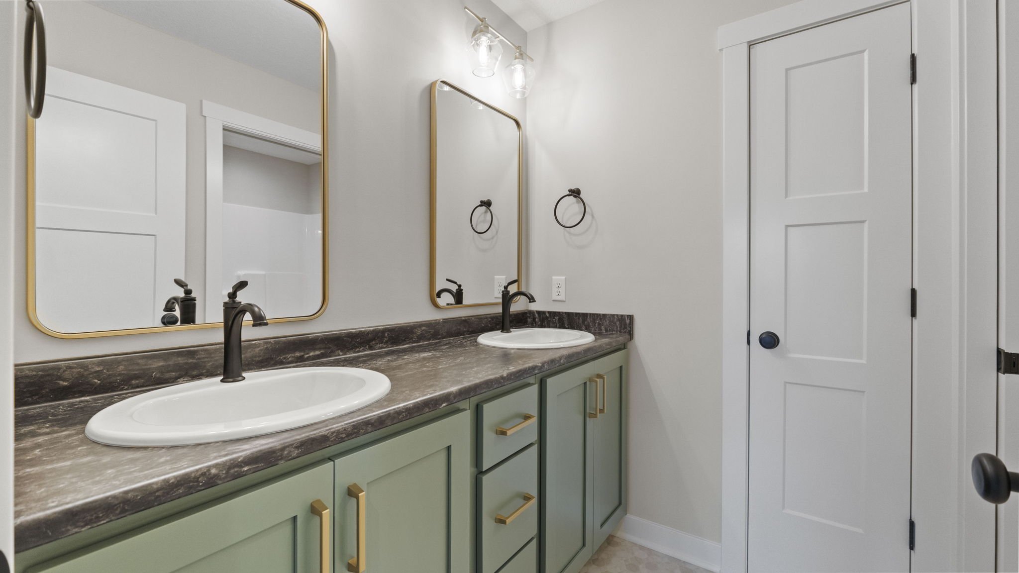 A bathroom vanity with two sinks, dark marble countertop, green cabinets with gold handles, two mirrors, and a white door.