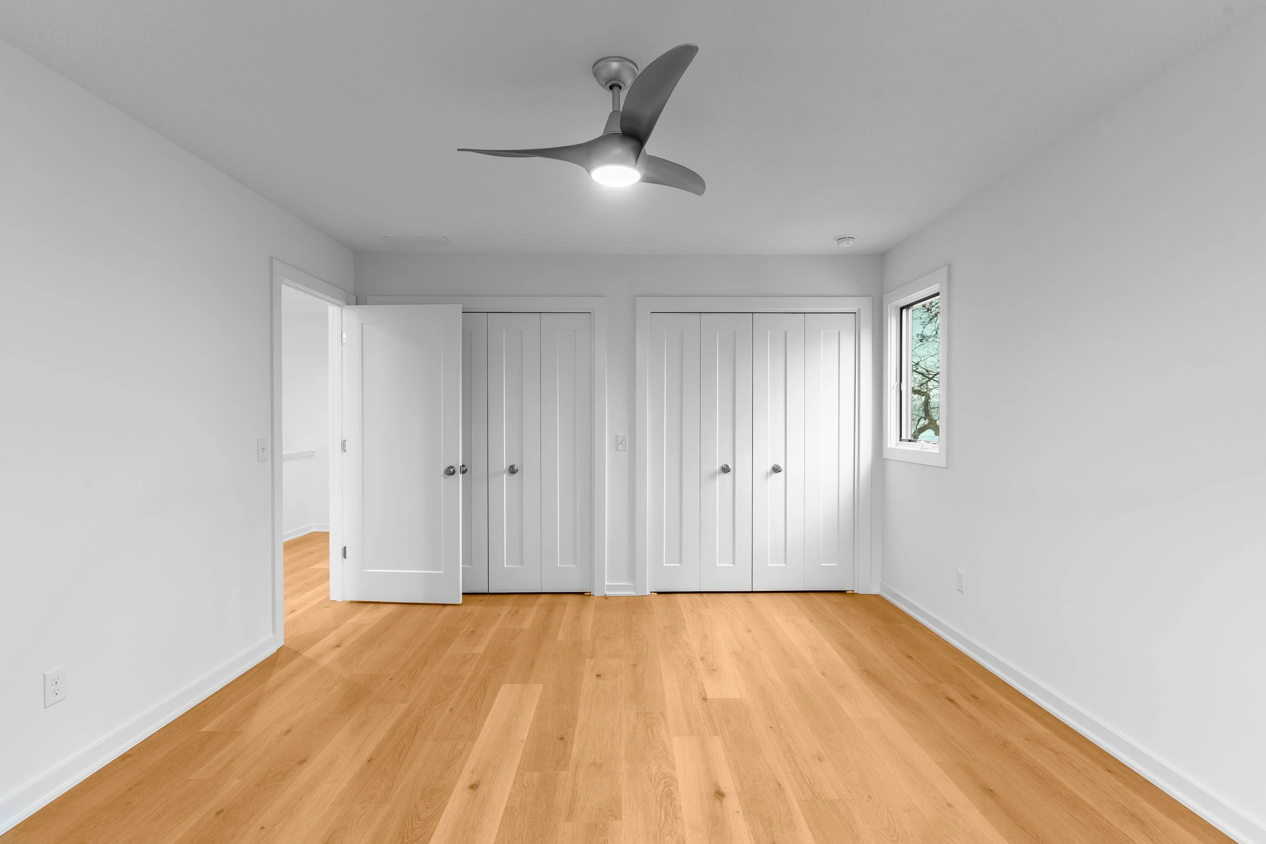 Empty bedroom with hardwood floors, white walls, two closets with white doors, one window, and a modern ceiling fan with light.