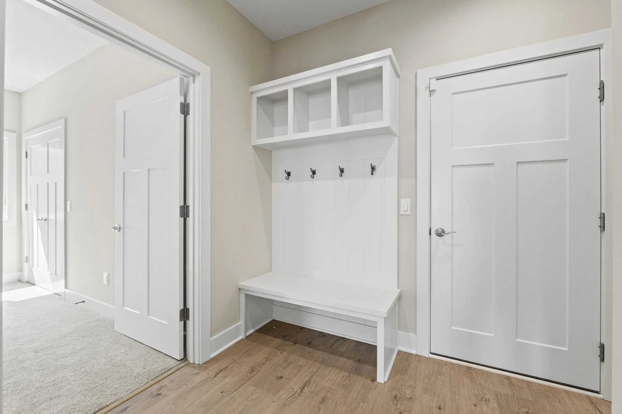 White entryway bench and wall hooks with storage cubby, located near plain white doors, in a room with beige walls and light wood flooring.