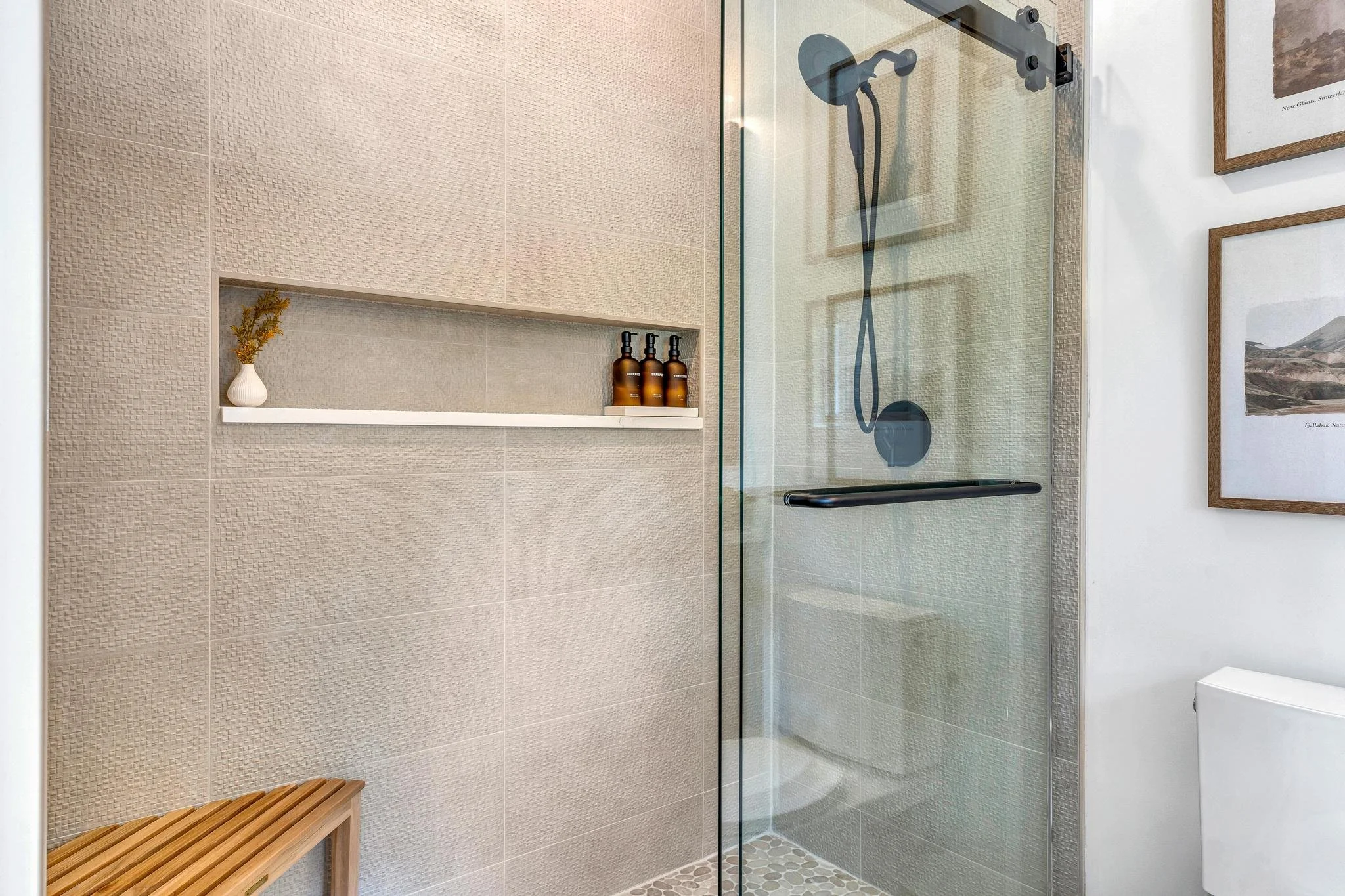 Modern walk-in shower with glass door, beige textured wall tiles, a built-in niche with small bottles and a vase, a wooden bench, and framed pictures on the white wall.