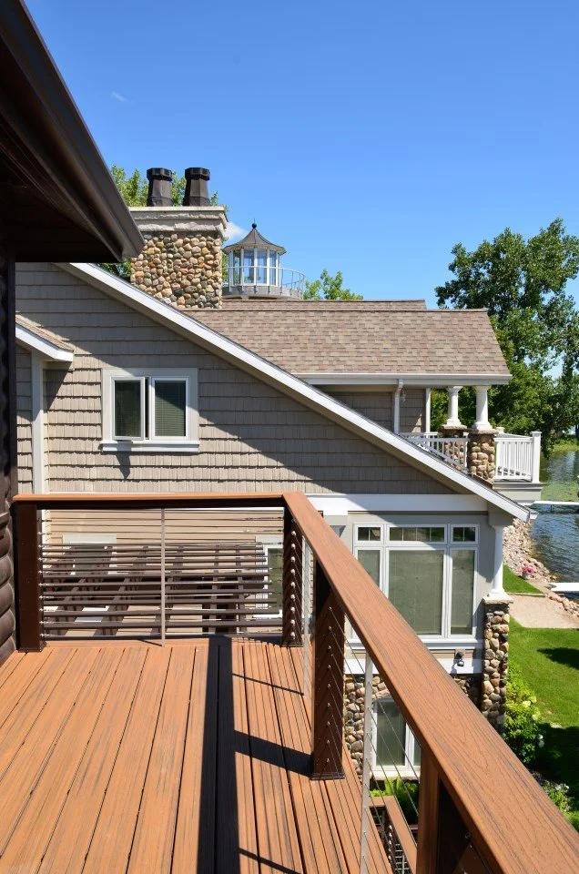 View of a house with a wooden deck, slate siding, and a stone chimney, overlooking a garden and a body of water in the background under a clear blue sky.