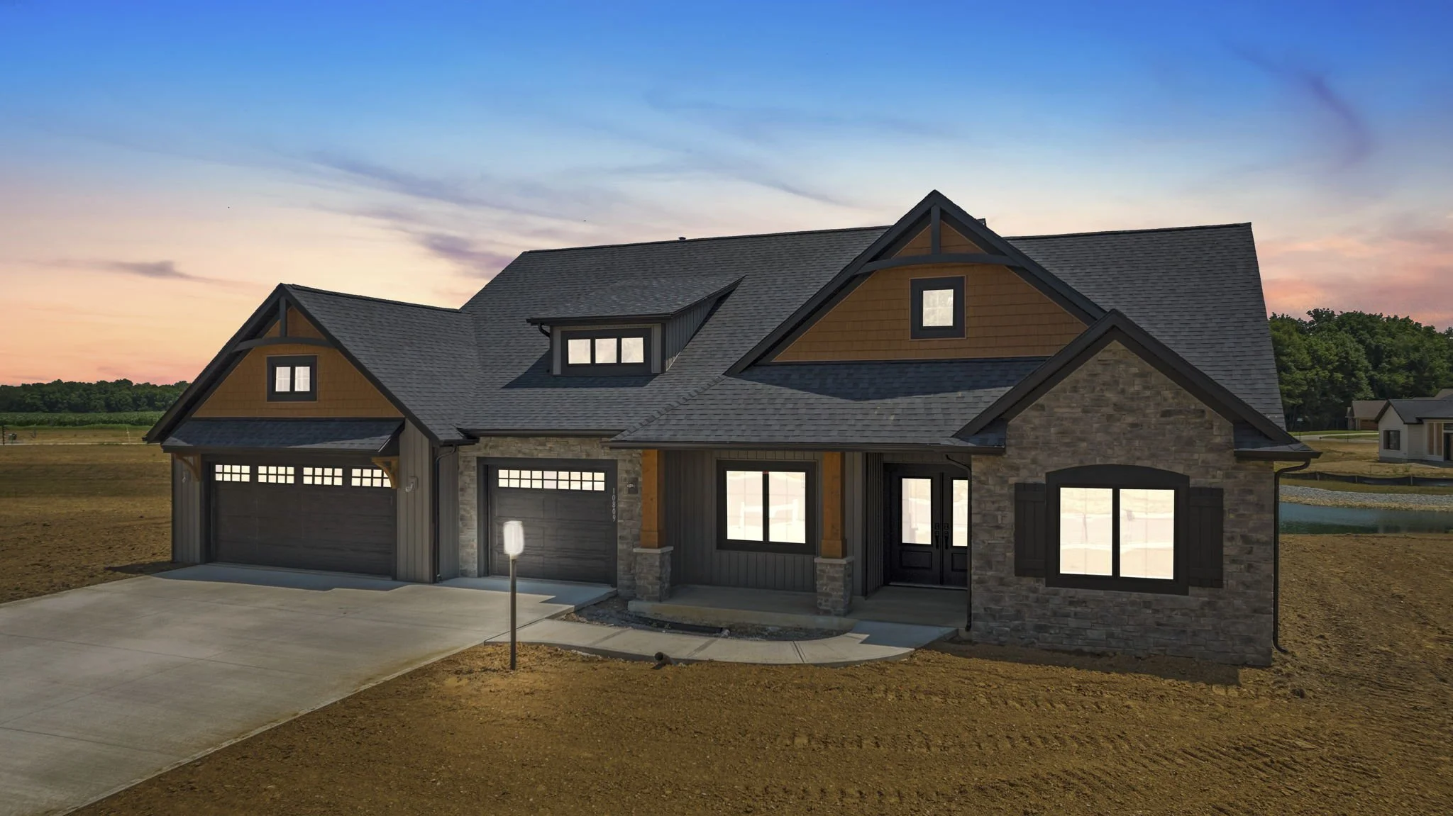 A two-story house with a dark gray roof, brick and wooden exterior, black window trims, and a three-car garage with black doors, surrounded by a dirt yard at sunset.