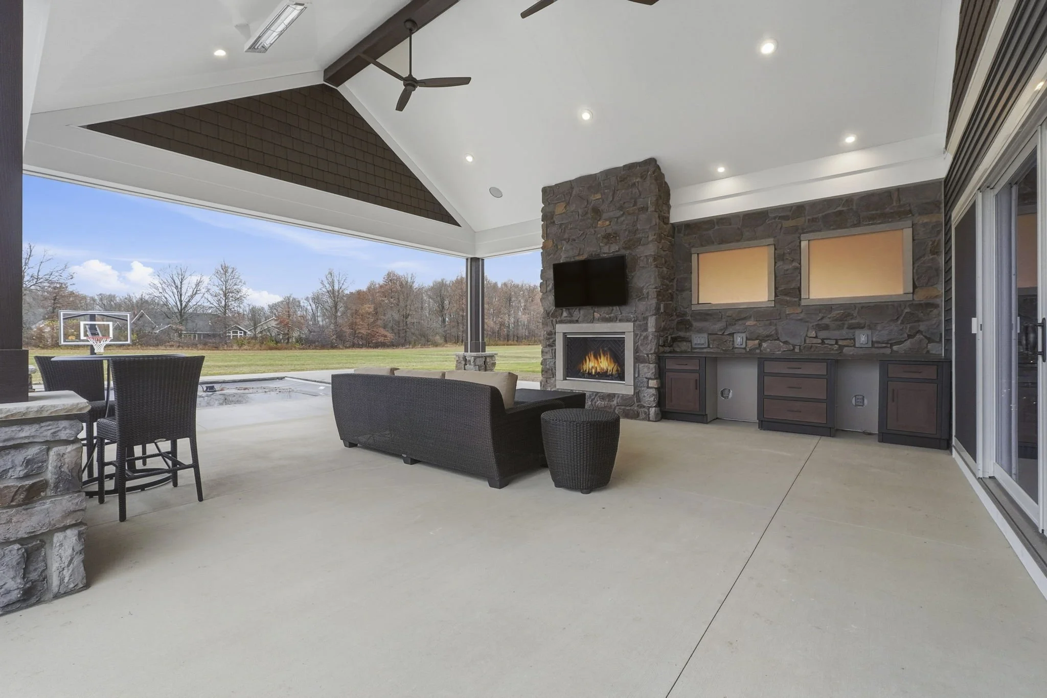 Covered outdoor patio with stone fireplace, wall-mounted TV, black wicker furniture, and view of grassy field with basketball hoop and trees in the background.