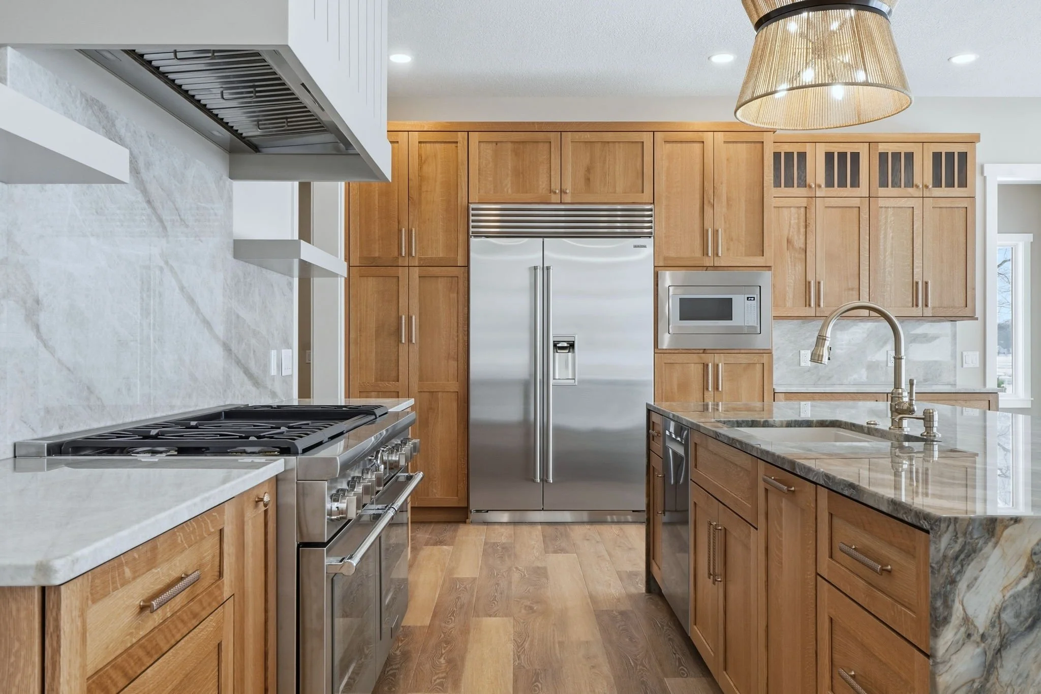 Modern kitchen with wooden cabinets, stainless steel refrigerator, microwave, and a marble countertop island with a sink and a brass faucet. Wooden flooring and a large window in the background.