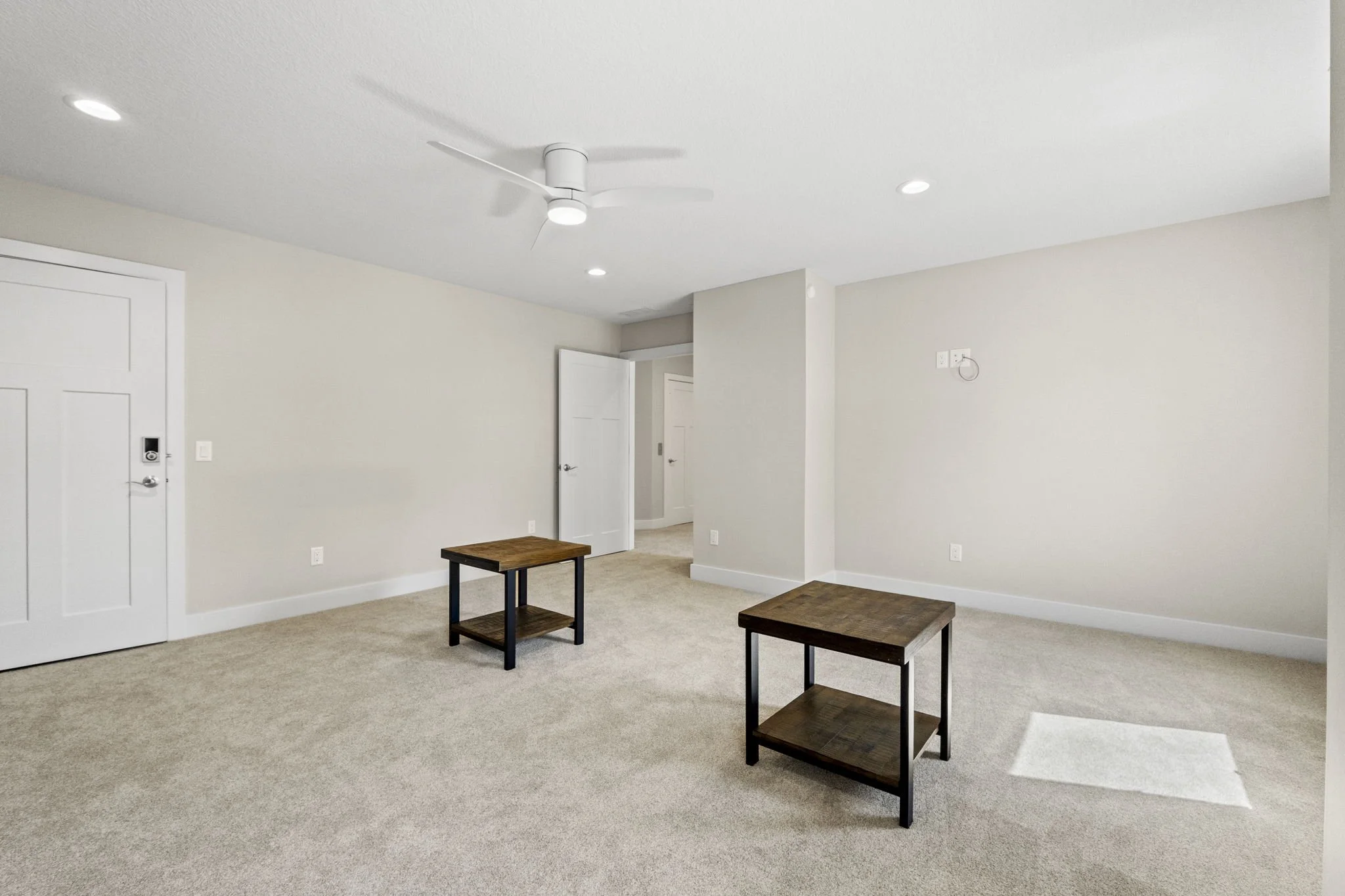 Empty living room with beige carpet, two small wooden tables, white ceiling fan, white walls, and recessed lighting.