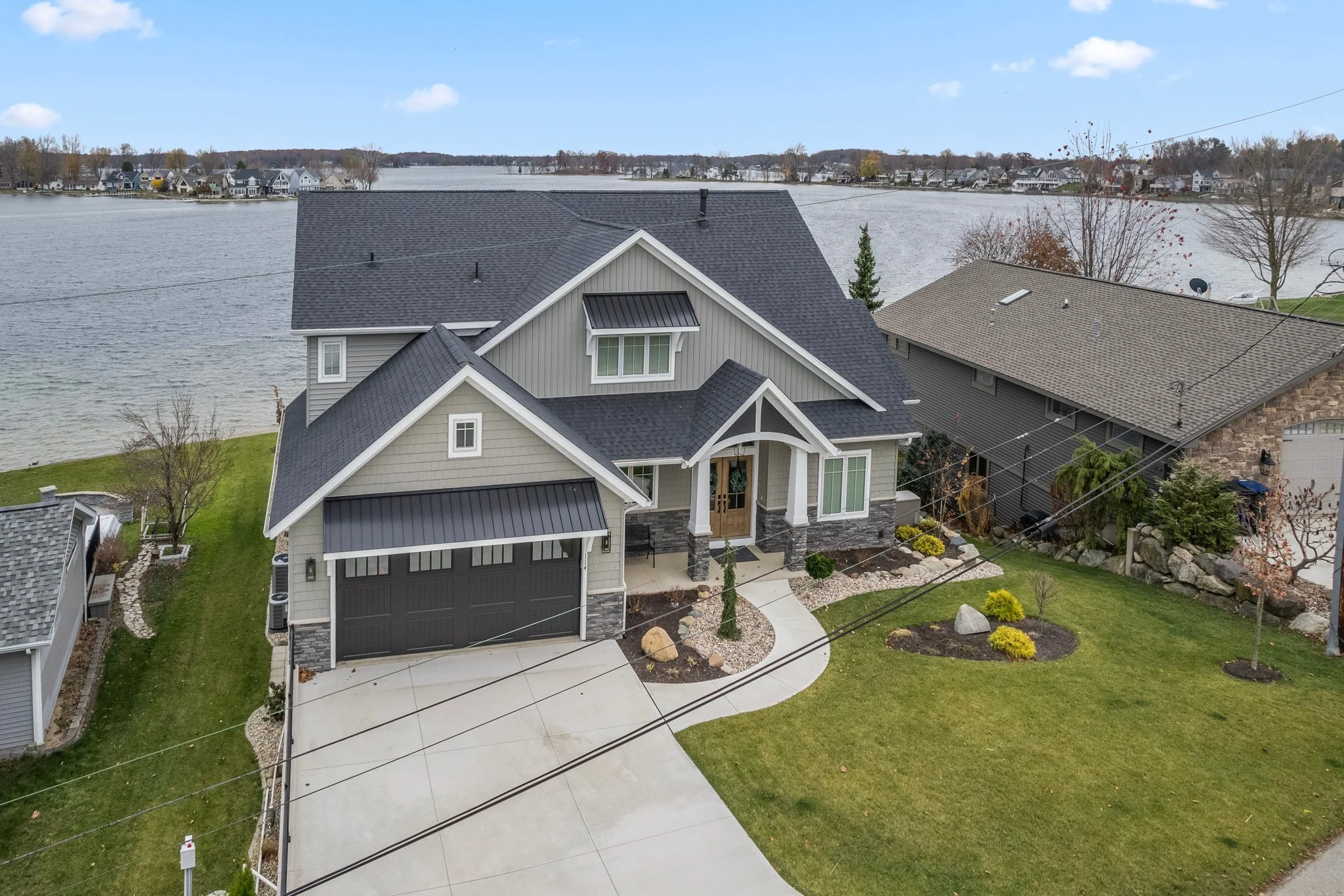 A modern two-story house with a gray and white exterior, black garage door, and a landscaped front yard, situated near a large body of water under a partly cloudy sky.