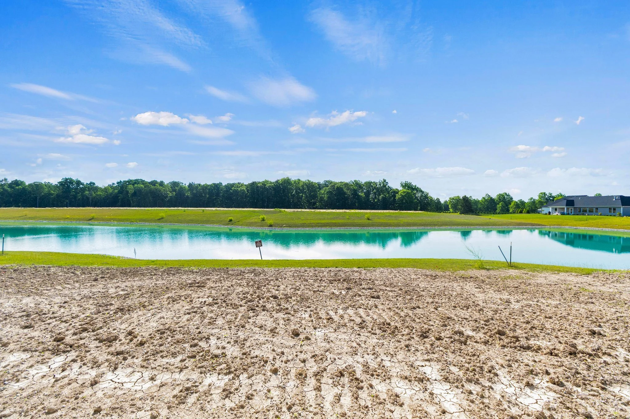 A small body of water, possibly a pond or reservoir, with a reflecting surface under a bright blue sky with scattered clouds, surrounded by grassy areas and a few tiny trees, with a house visible in the distance.
