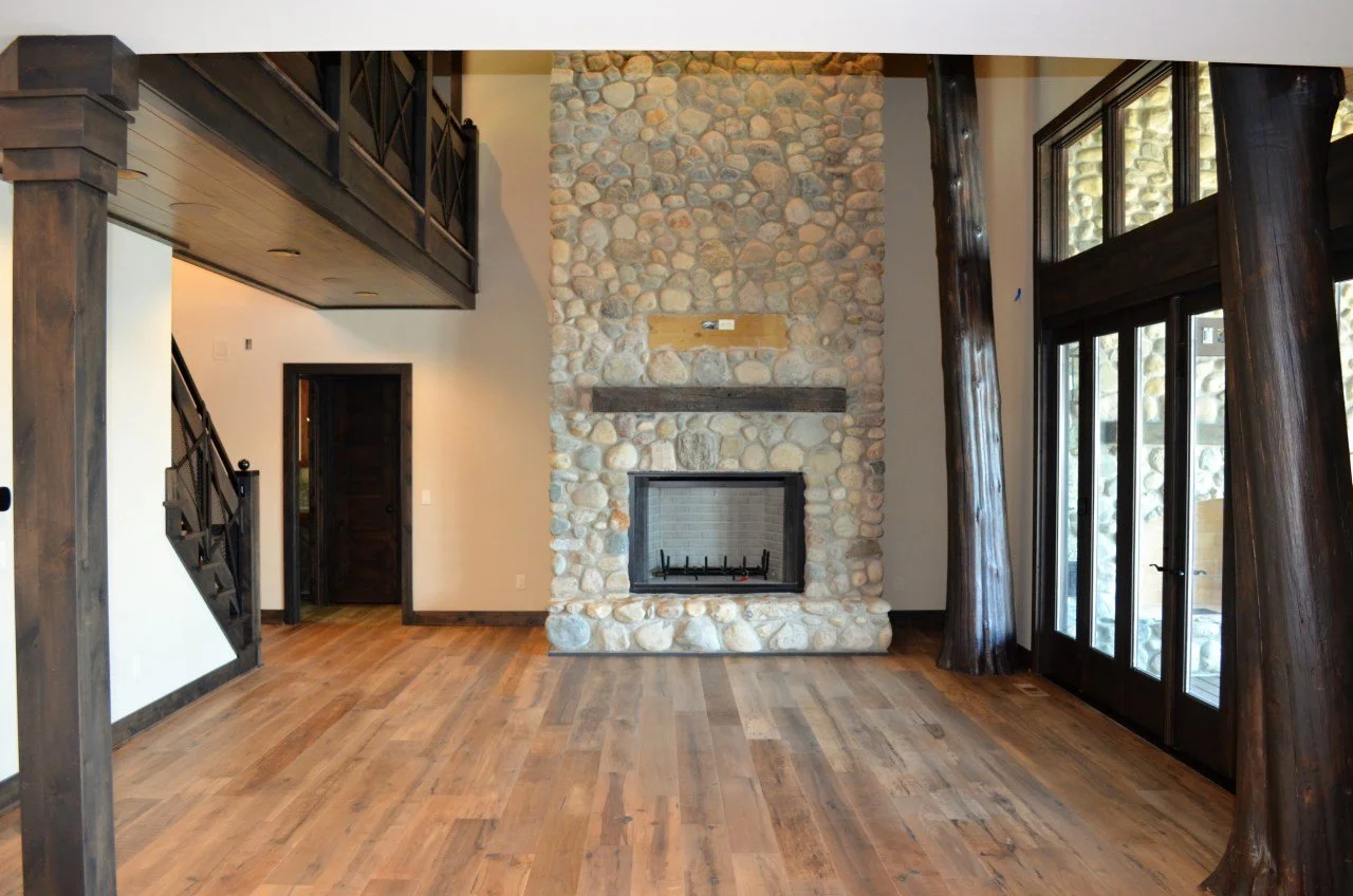 Living room with a stone fireplace, wooden floors, dark wood beams, and large glass doors.