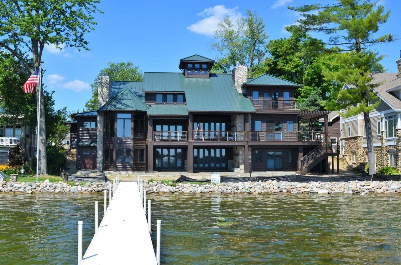 A large, modern waterfront house with dark wood siding and multiple decks, situated along a body of water with a dock extending into the water, surrounded by trees and neighboring houses.