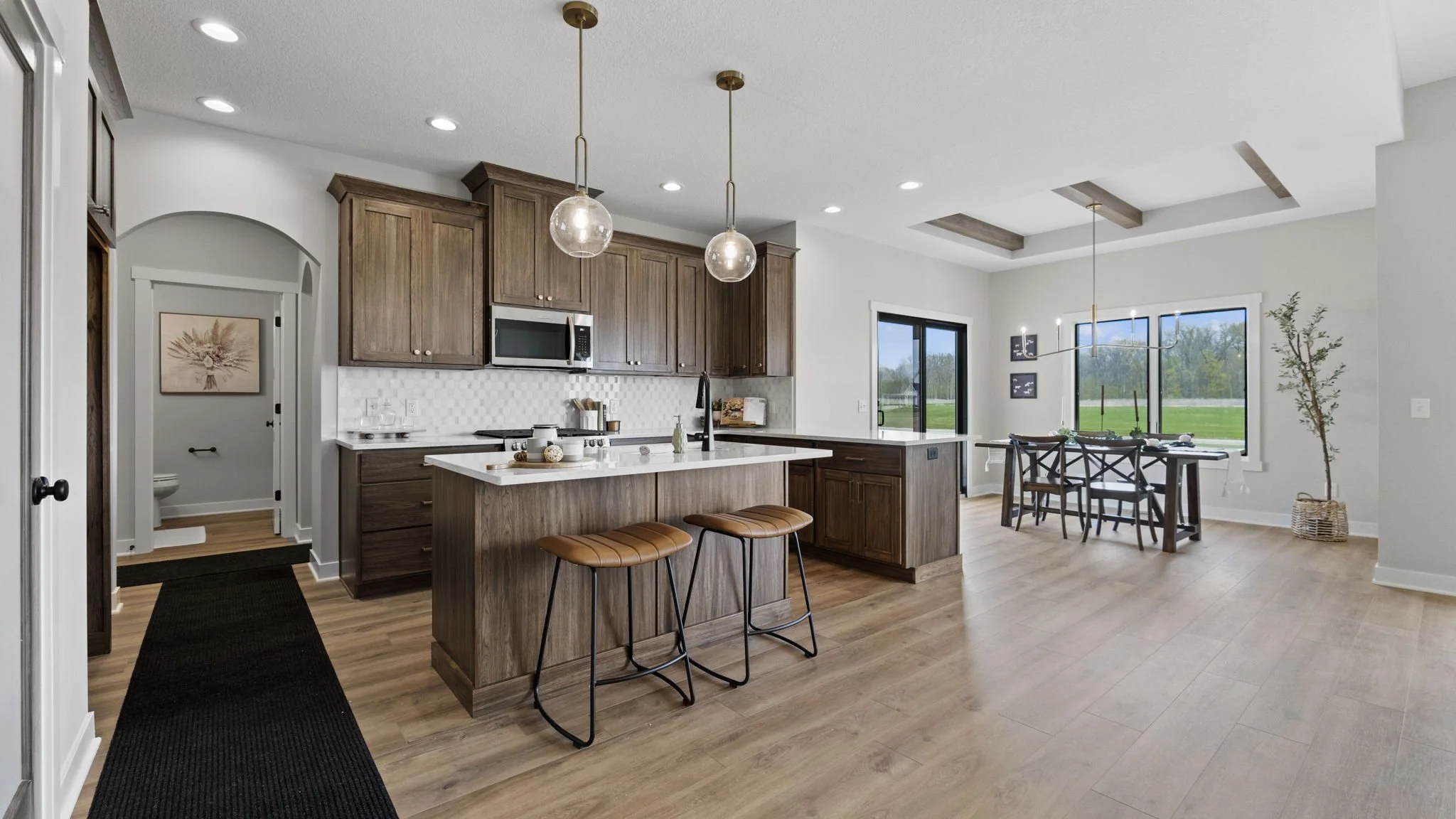 Open-concept kitchen with wooden cabinets, white island, two pendant lights, and a dining area with large windows overlooking a green field.