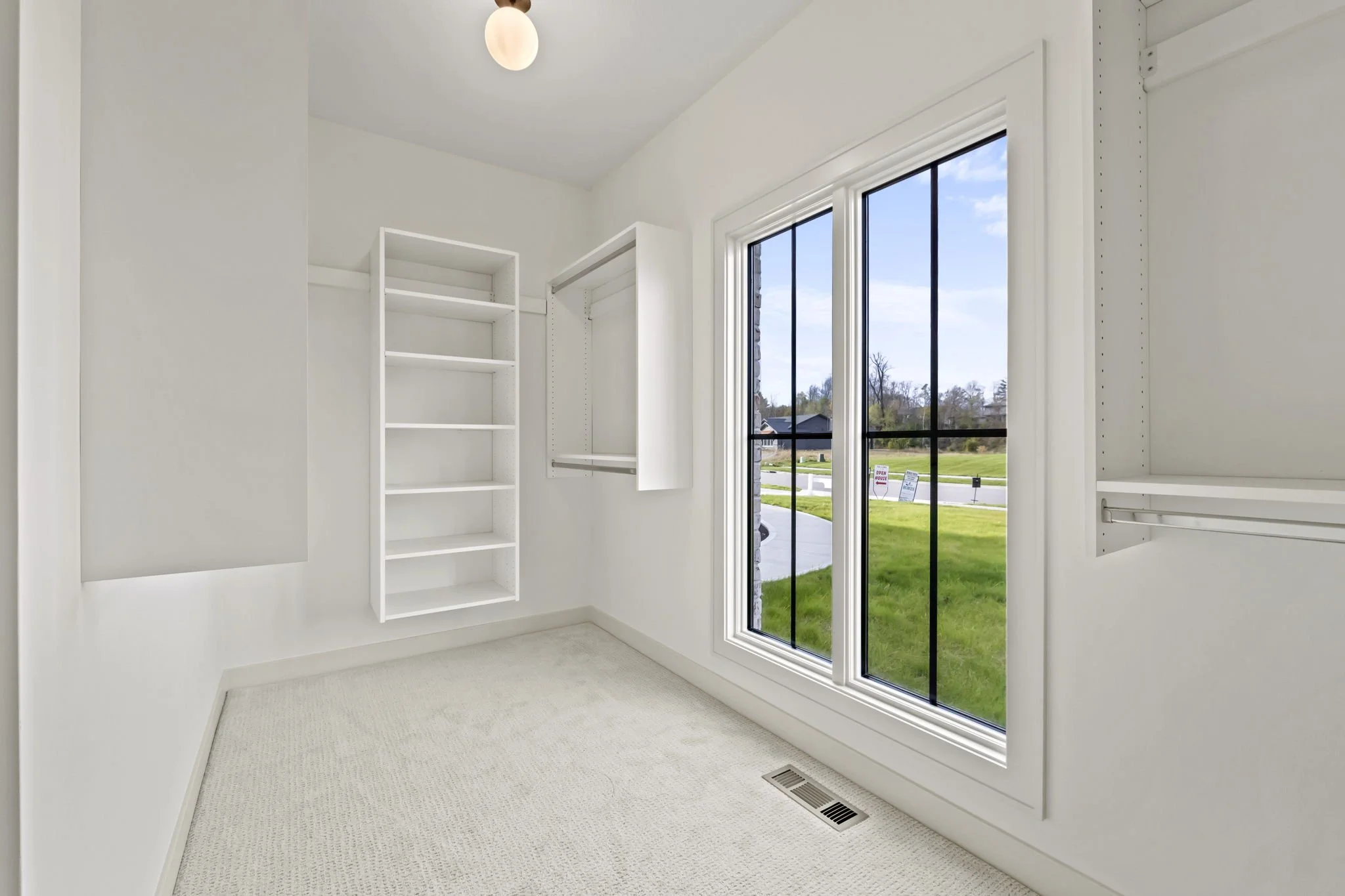 Empty walk-in closet with white built-in shelves and hanging rods, beige carpet, and large window showing a suburban street with green grass and trees.