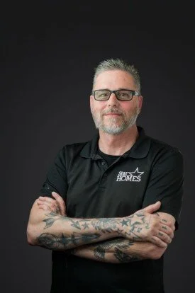 Man with gray hair and beard, wearing glasses and a black polo shirt with a logo, standing with arms crossed against a dark background.