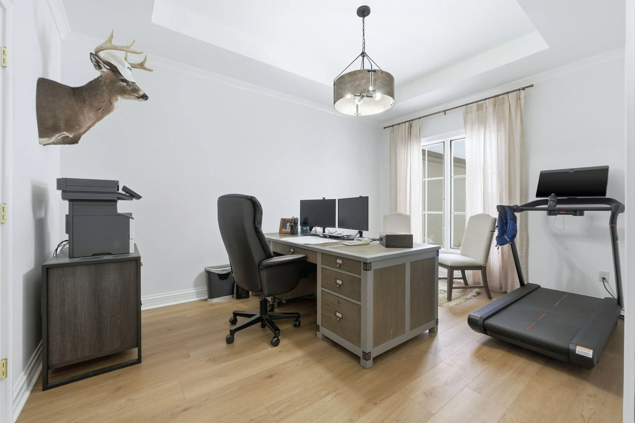 Home office with desk, two monitors, office chair, treadmill, paper shredder, and mounted deer head with a cap.