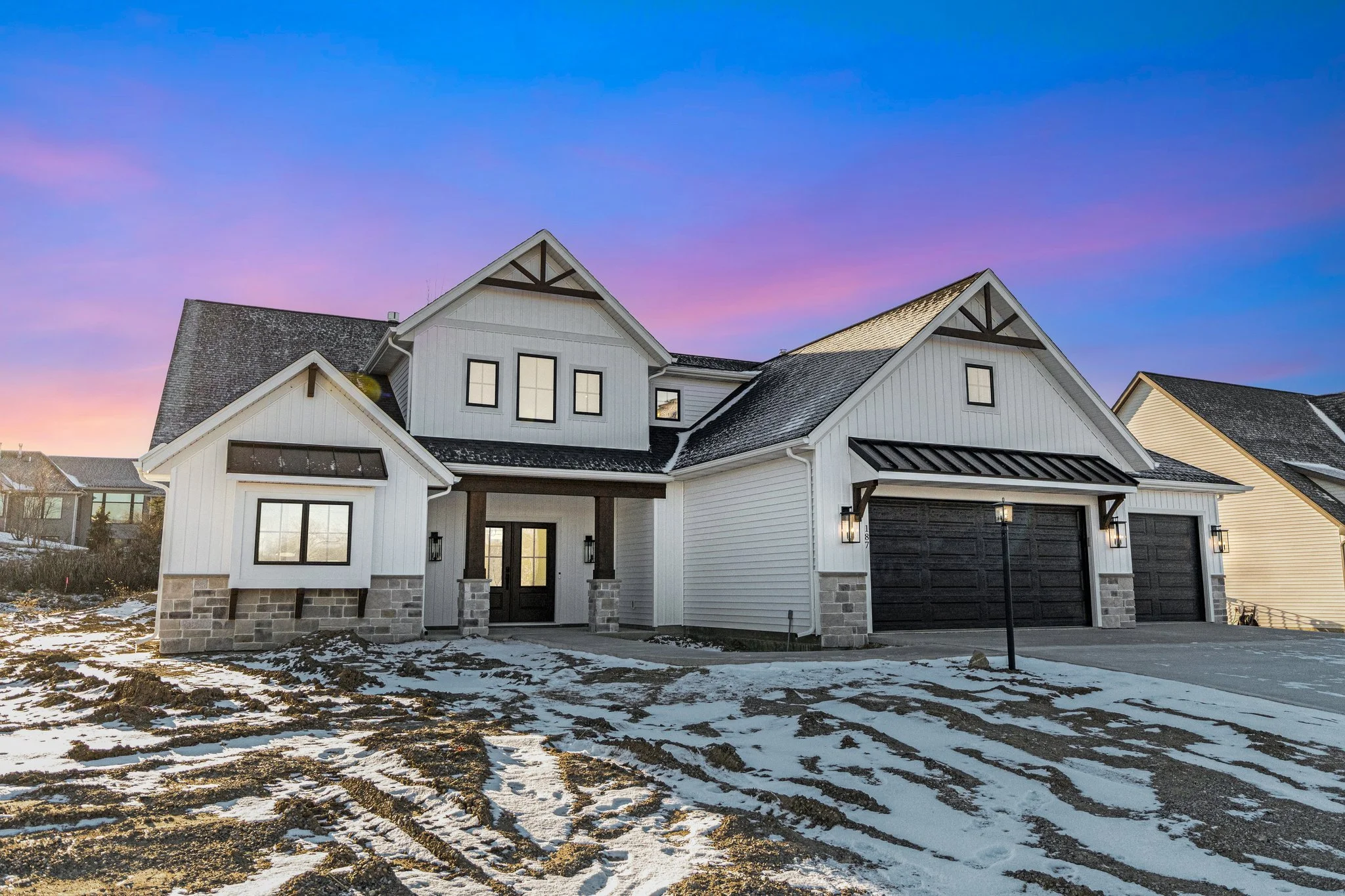 Modern two-story house with white siding, black garage doors, and stone accents. Snowy front yard with tire tracks, sunset sky with pink and purple hues.