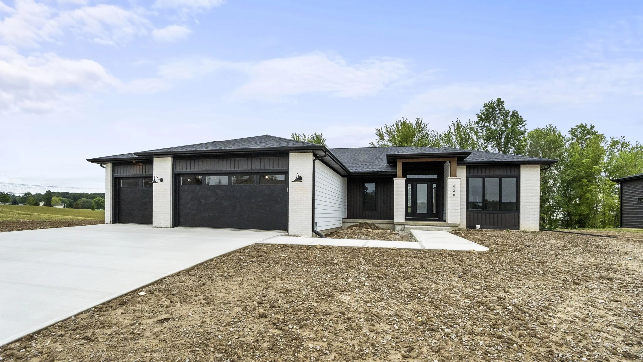 Newly constructed modern house with black and white exterior, black garage doors, and a concrete driveway, surrounded by a dirt yard and green trees in the background.