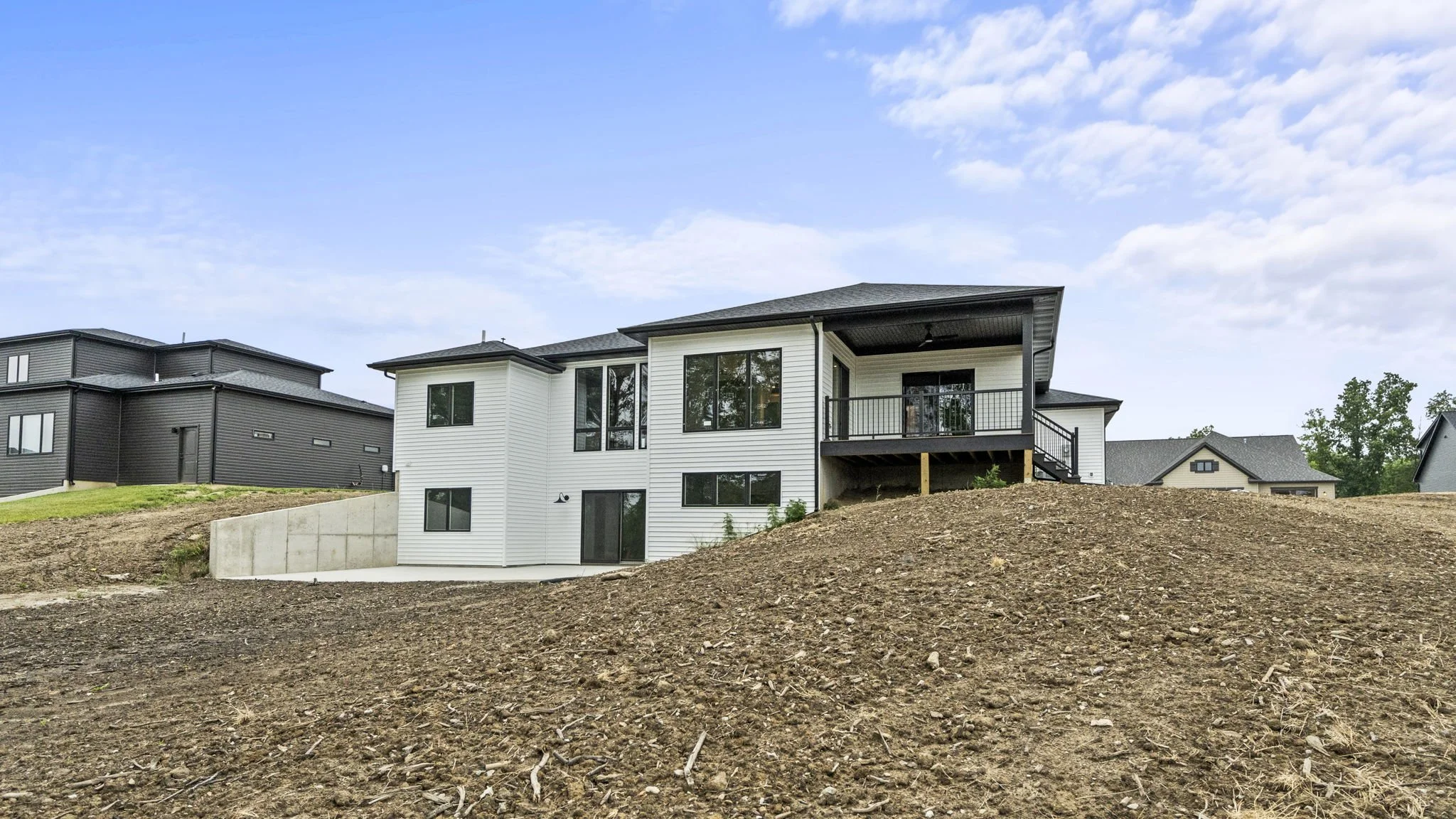 A modern two-story house with white siding, large windows, and a balcony on the upper level, built on a sloped lot with exposed dirt in the foreground and neighboring houses in the background under a partly cloudy sky.