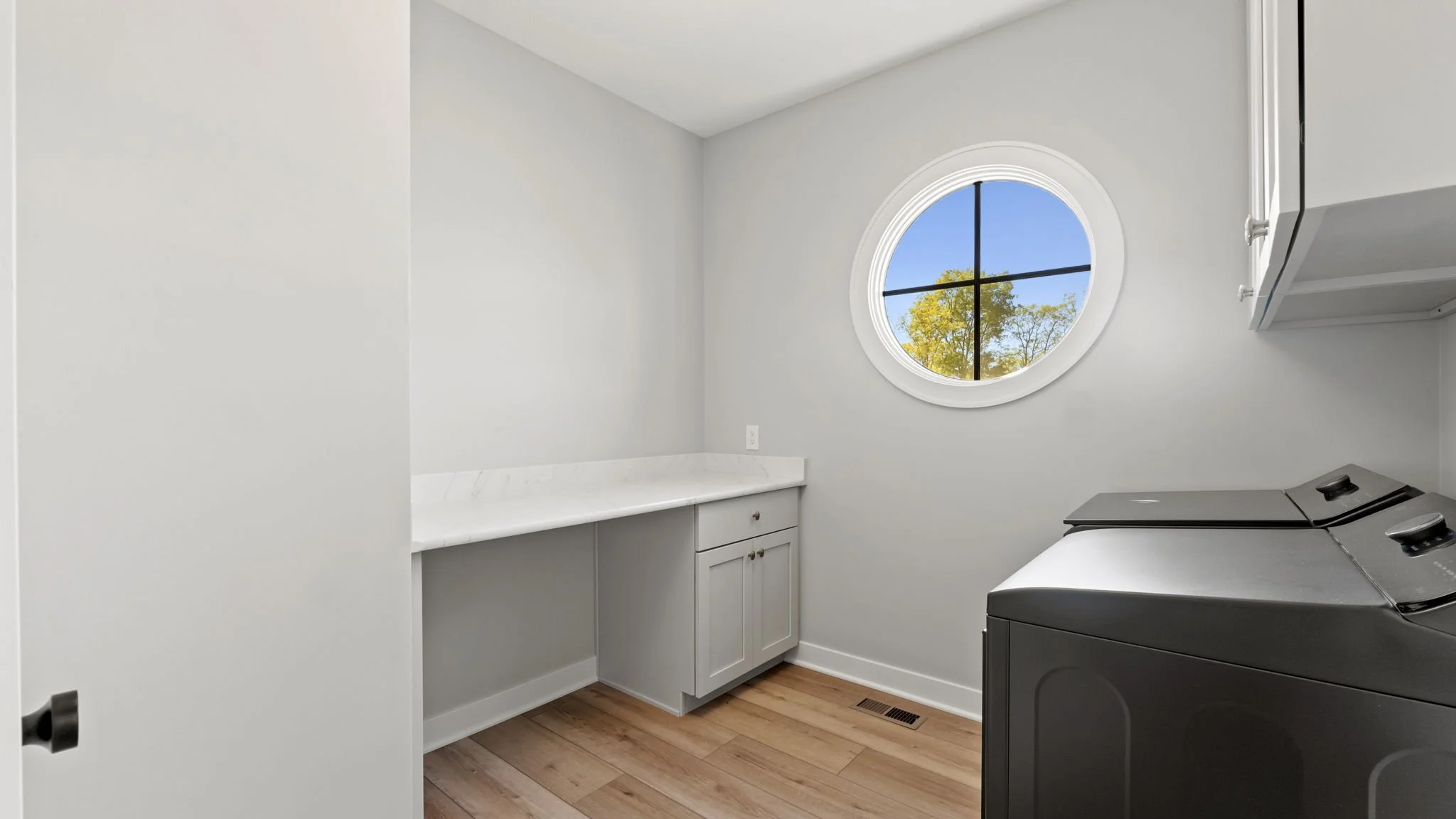 A laundry room with white walls, a round window showing blue sky and trees, a white countertop, gray cabinets, and a black washing machine.