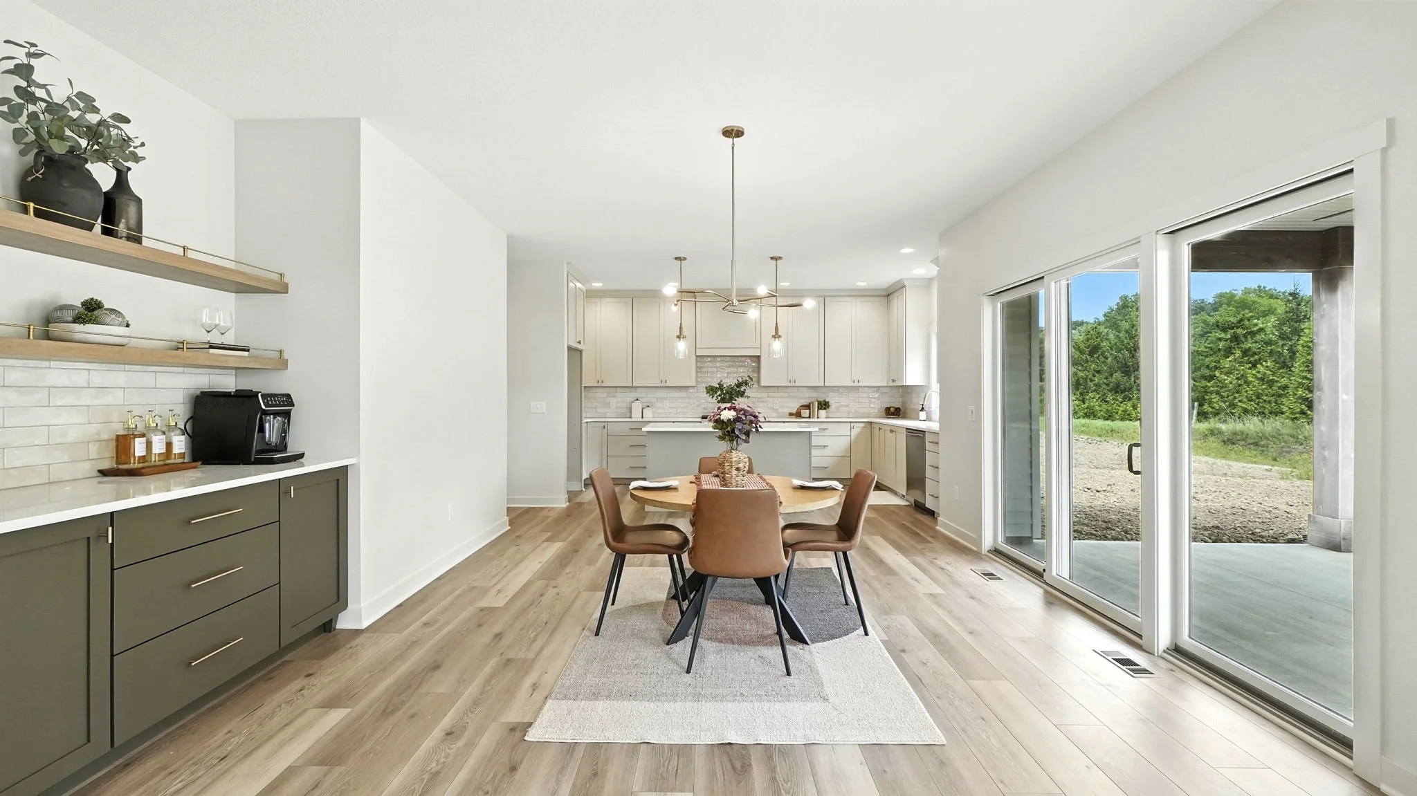 Modern kitchen and dining area with a round table, four brown chairs, a vase of pink flowers, hardwood flooring, sliding glass doors showing outdoor greenery, white cabinetry, and pendant lights.