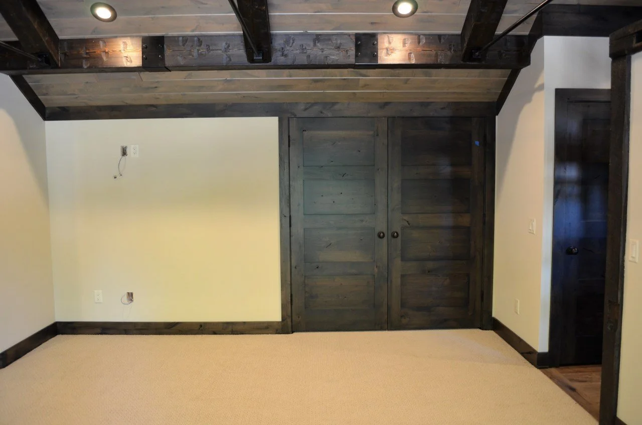 Empty room with beige carpet, cream-colored walls, dark wood trim, and a wooden ceiling with black exposed beams. There are electrical outlets and missing light fixtures on the walls. A large dark wood sliding door closet is in the middle of the wall