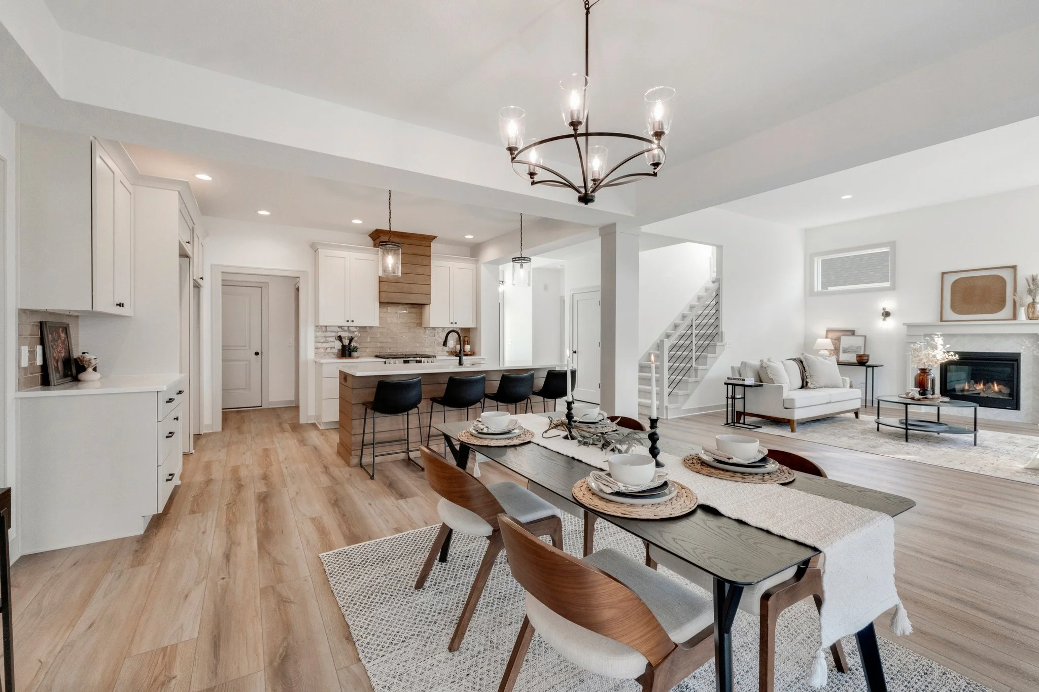 Open-concept living room and kitchen with wood flooring, white cabinets, a central island with black stools, a dining table with place settings, a fireplace with a mantel, and minimalist decor with neutral tones.