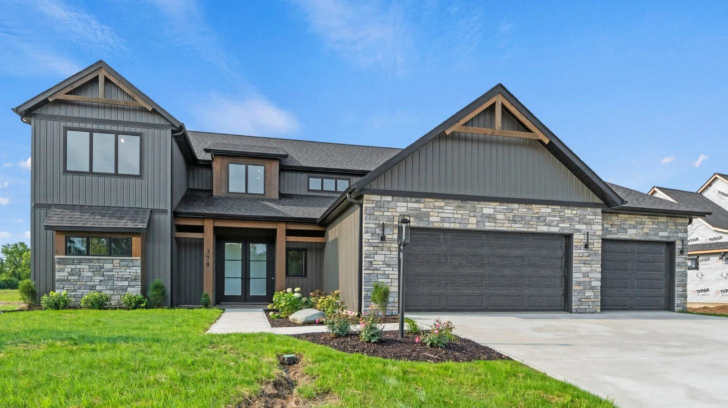 Modern two-story house with black and gray siding, stone accents, and a three-car garage, surrounded by a well-maintained lawn and garden with blooming flowers, under a blue sky.
