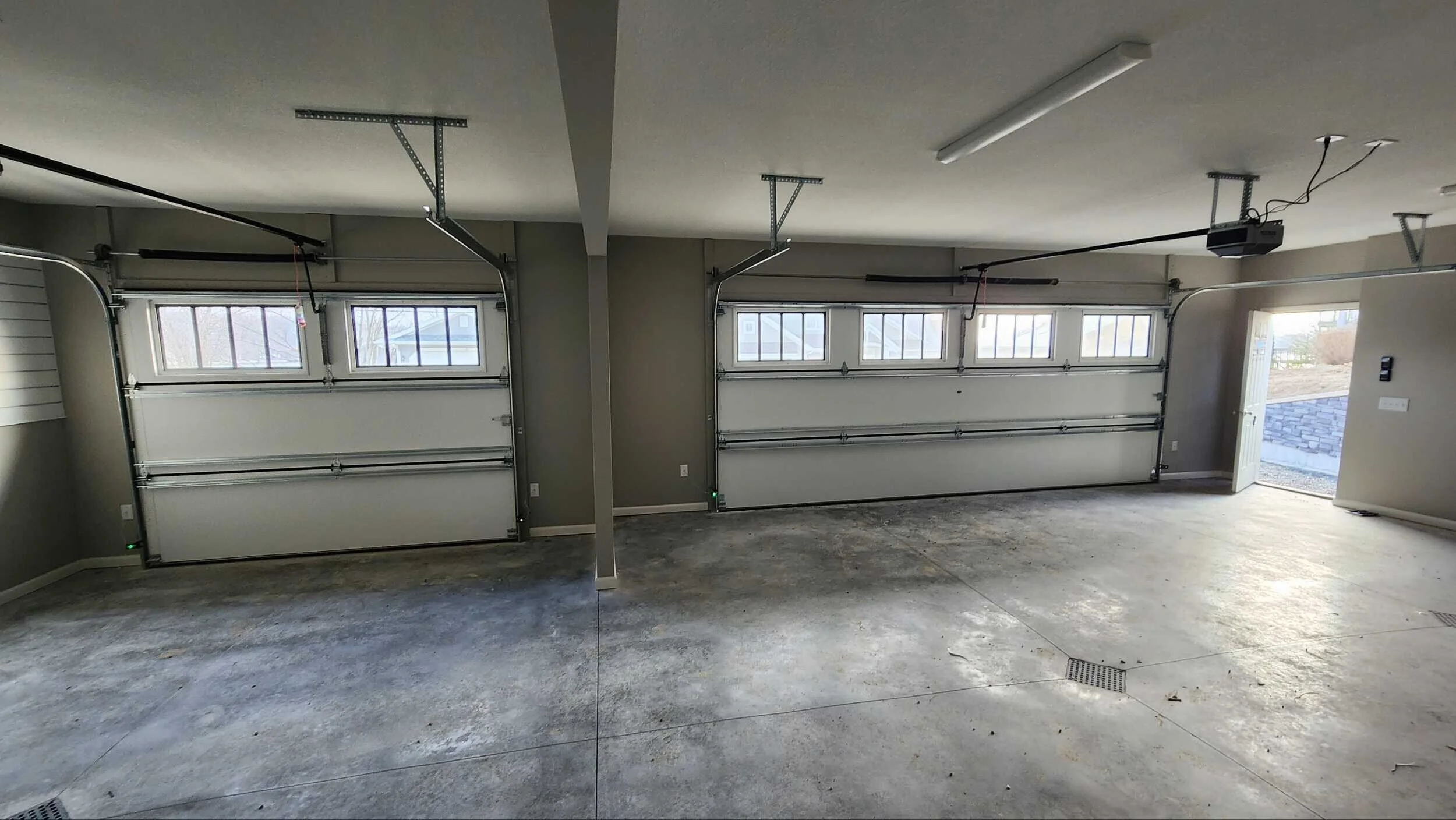 Empty two-car garage with concrete floor, two closed garage doors with windows, and an open door leading outside. Ceiling has a fluorescent light and garage automation system.