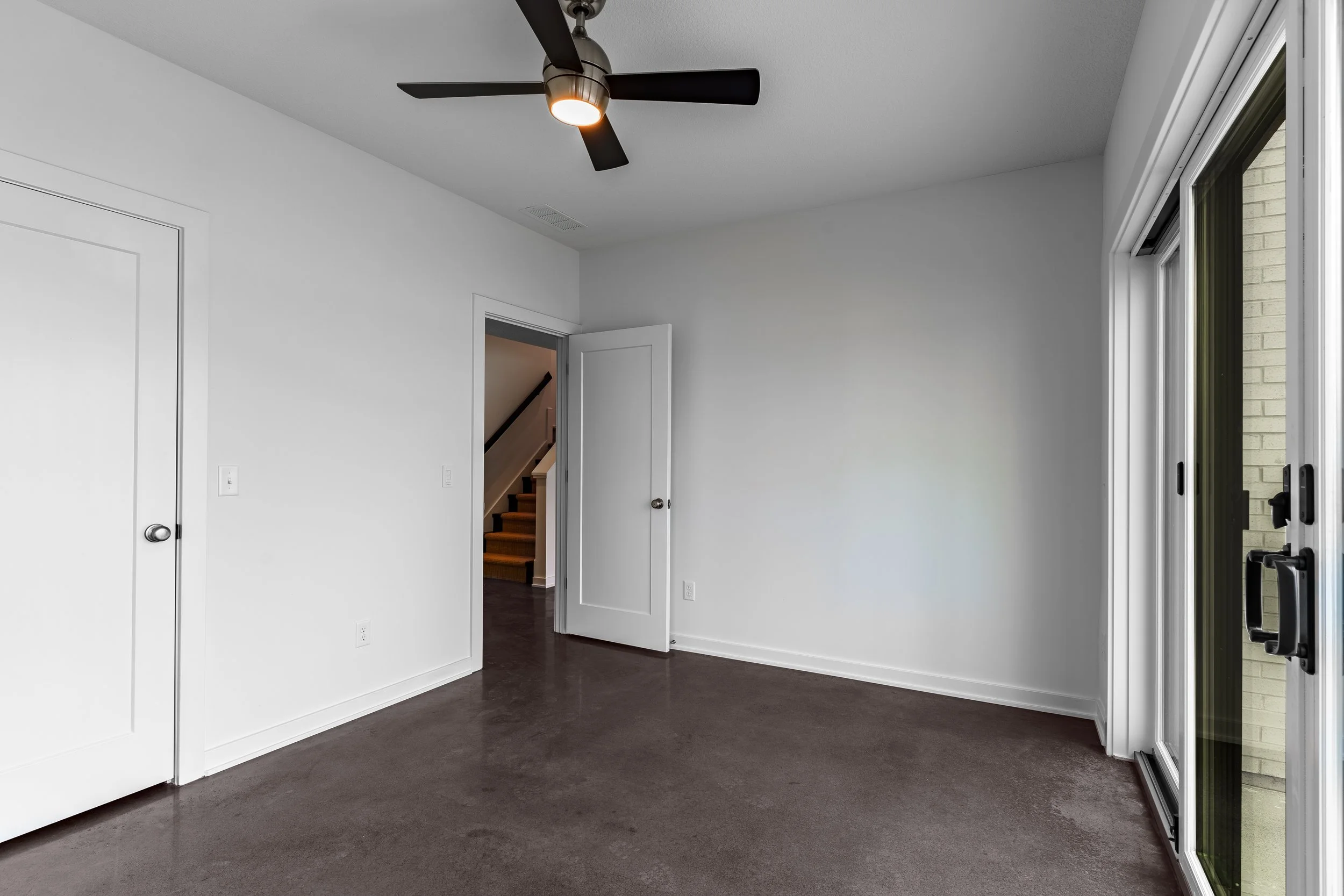 Empty room with white walls, dark brown carpet, sliding glass door, ceiling fan, and hallway with stairs.