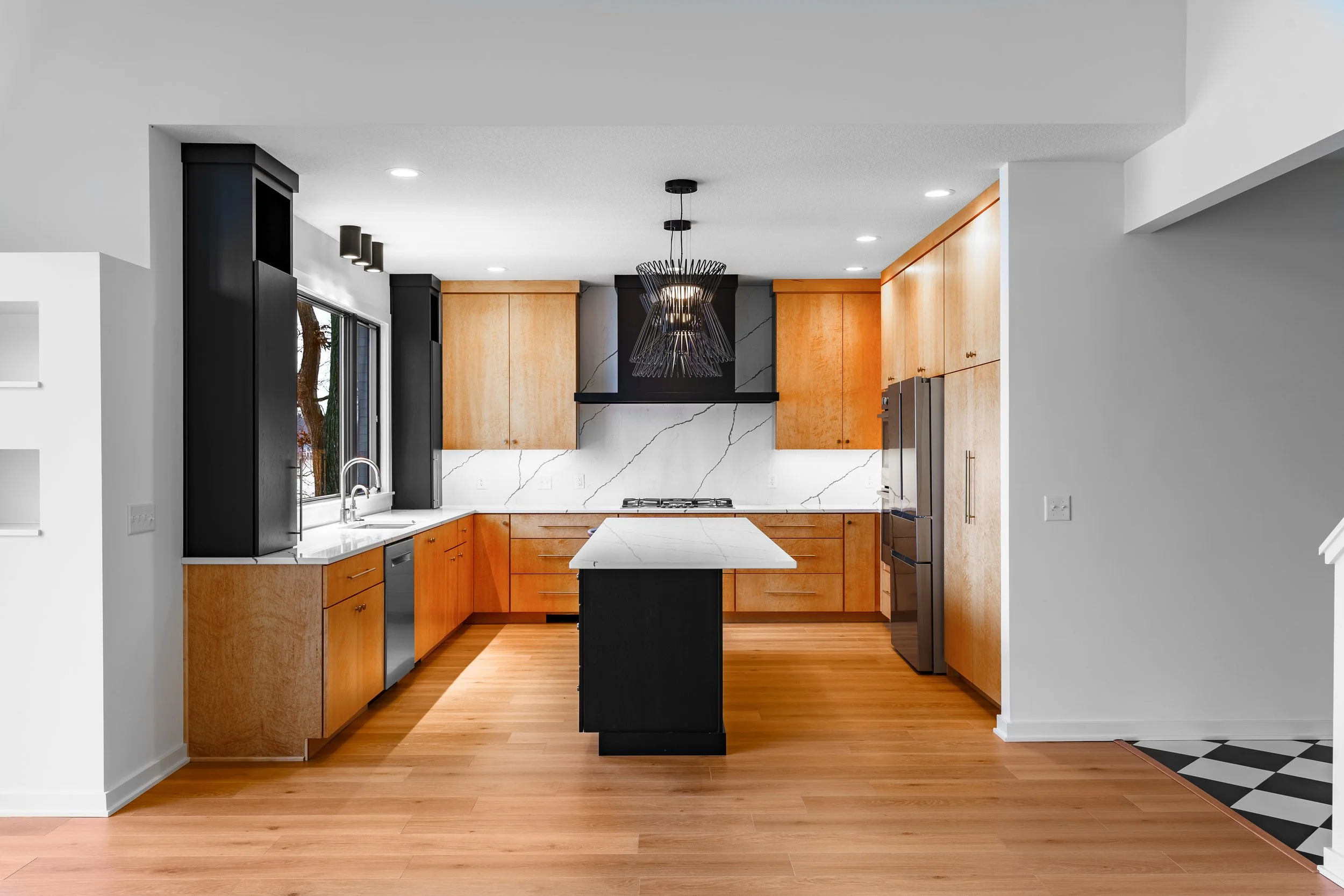 Modern kitchen with wooden cabinets, white marble countertops and backsplash, black accents, stainless steel appliances, and a central island with a black base and white marble top, hardwood floors, and a black artistic pendant light.
