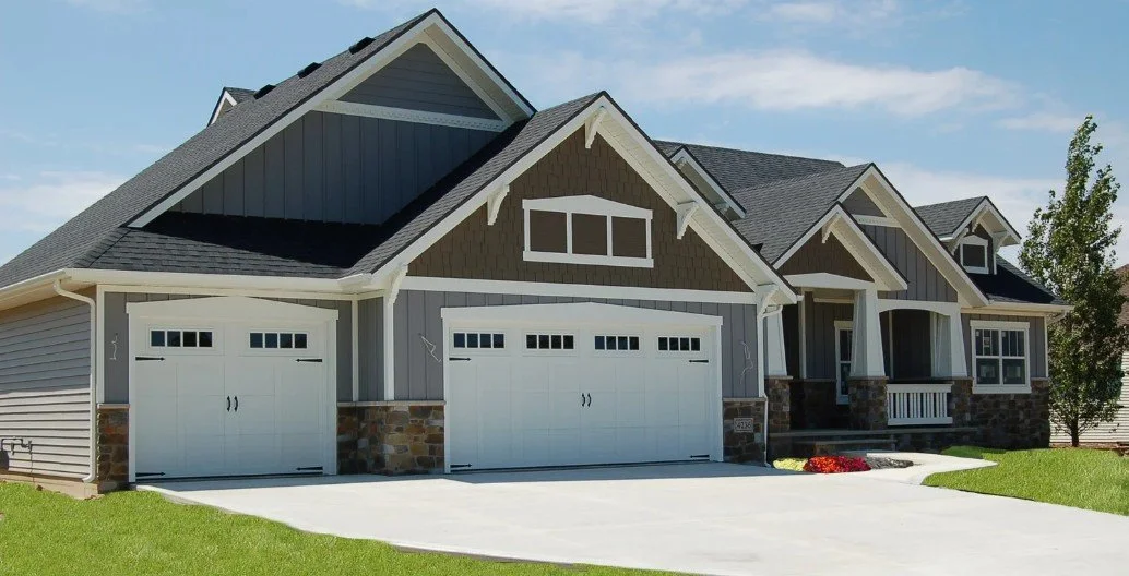 Front view of a modern suburban house with a double garage, gray siding, and a stone foundation, with a well-maintained lawn and a clear blue sky.