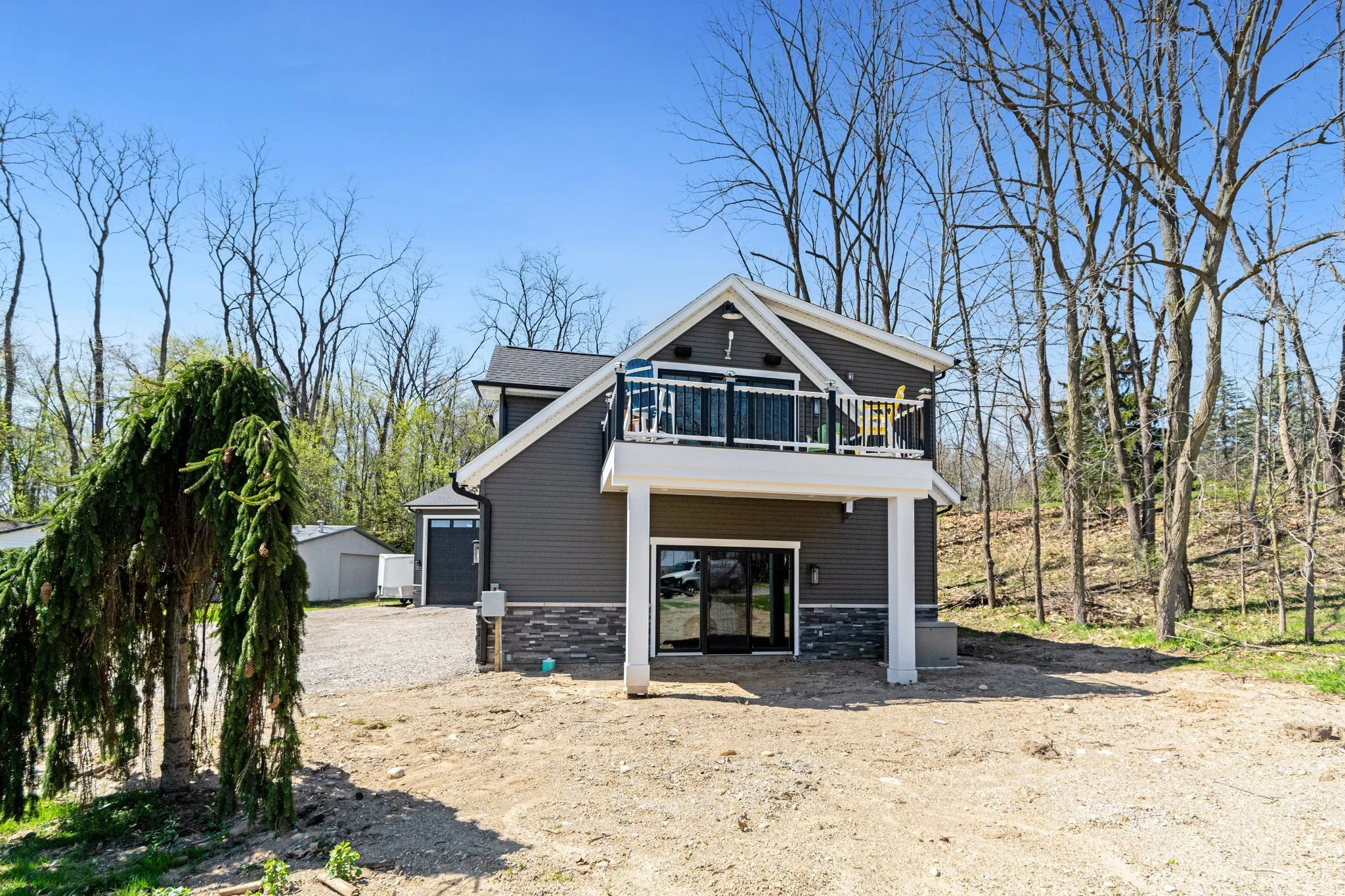 A modern two-story house with dark gray siding, white trim, a slate gray roof, and a small upper balcony with black railing, situated in a partly wooded area with bare trees, during a sunny day.
