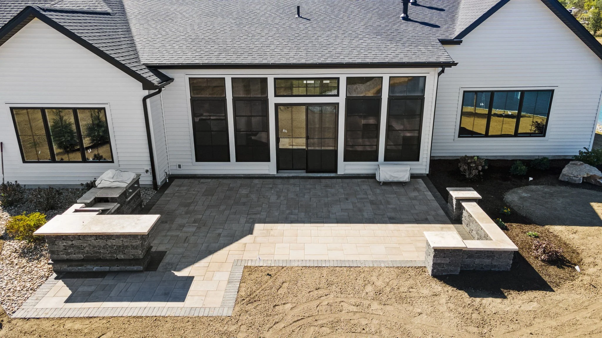 A newly constructed backyard patio with a combination of gray and beige pavers, separated by a decorative border. The patio is flanked by two stone benches with matching tiled tops, one on the left and one on the right. There's a built-in grill area 